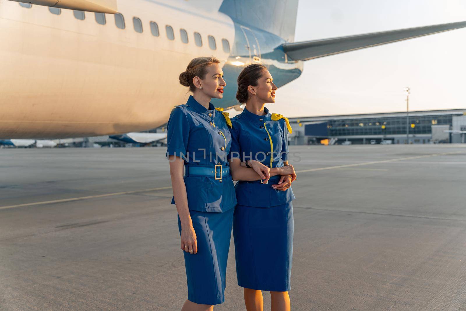 Two beautiful stewardesses in uniform standing near the plane and looking to the side