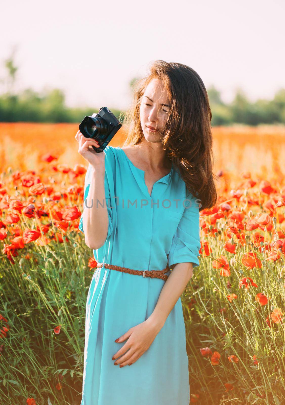 Beautiful young woman standing with photo camera in red poppies flower meadow.