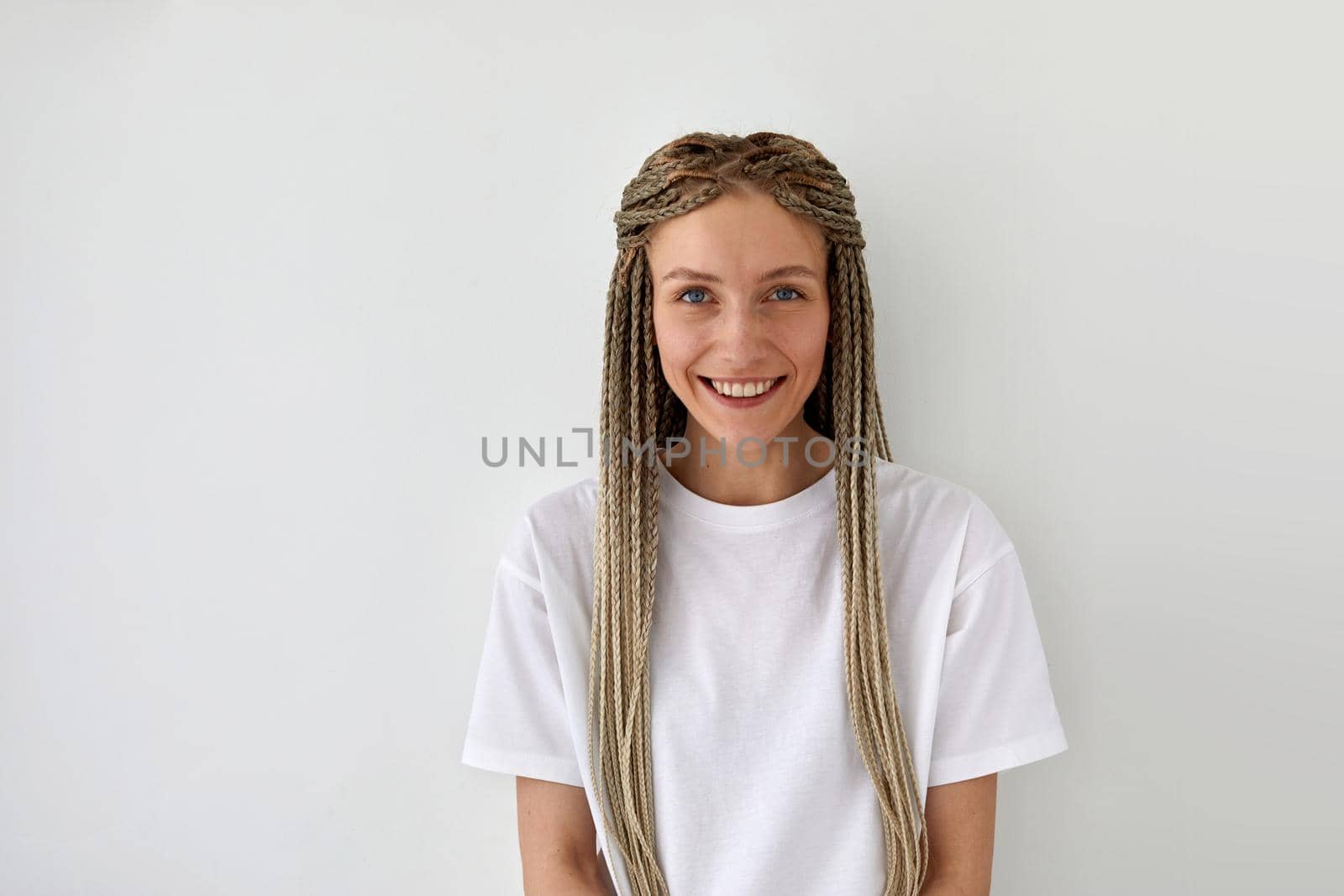 Cheerful attractive female with braids wearing casual white shirt standing on light background and looking at camera