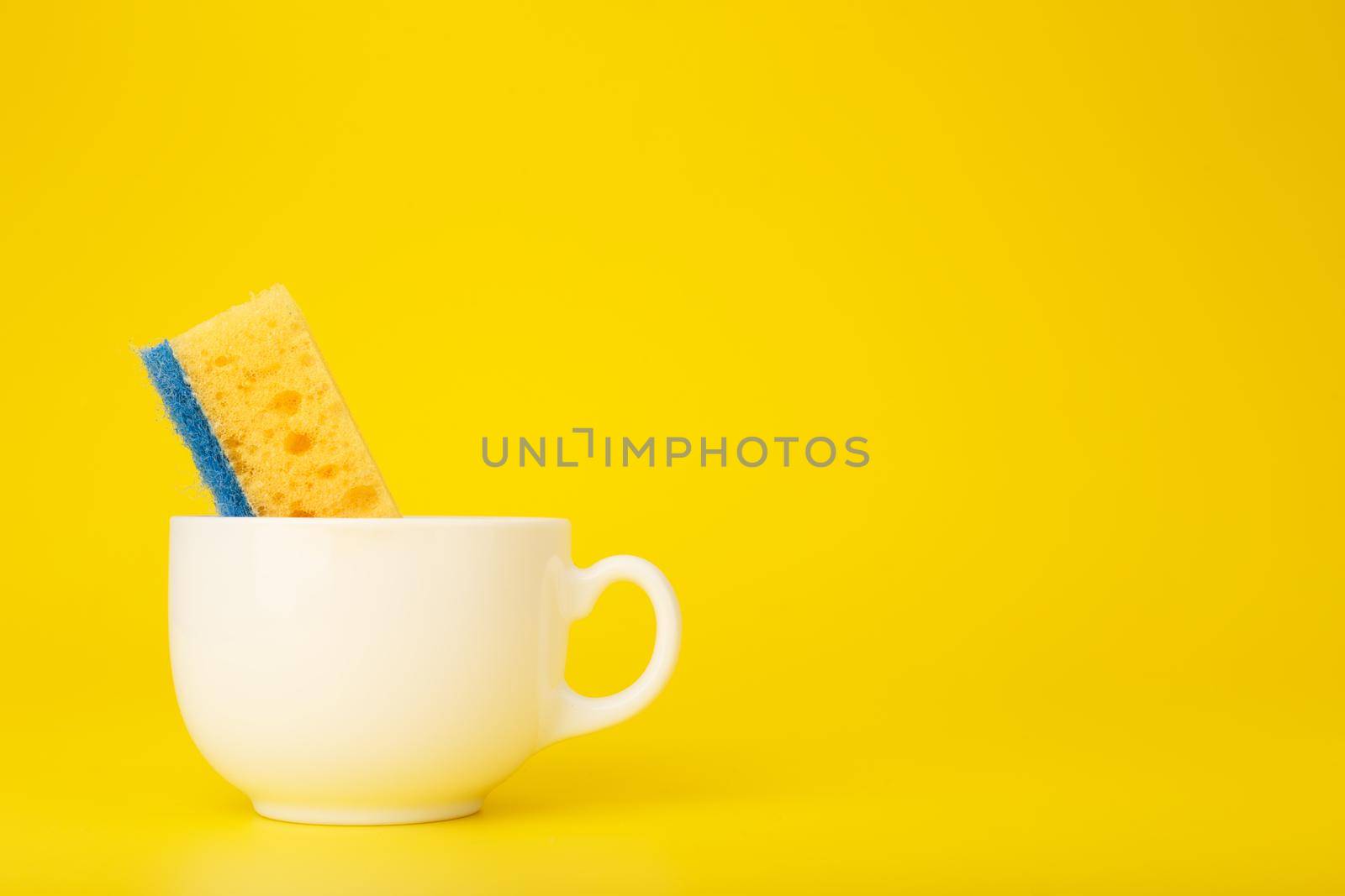 Creative, minimal dishwashing concept. Simple composition with yellow cleaning kitchen sponge in white ceramic cup on yellow background with copy space.