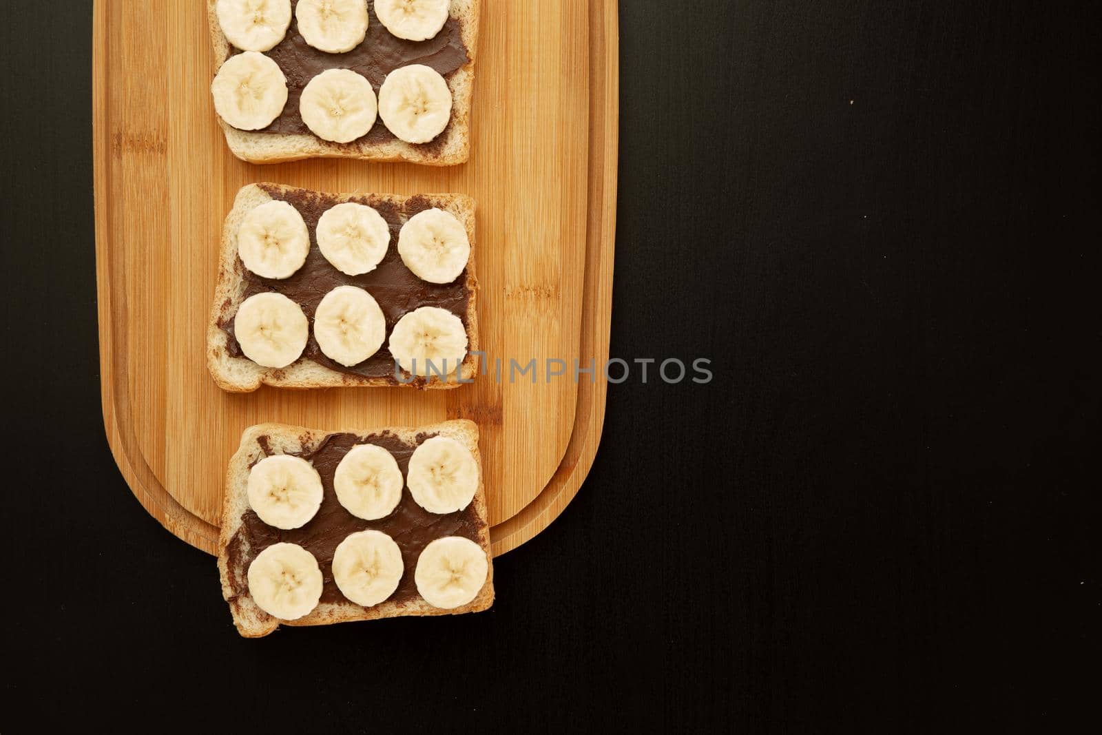 Three banana white bread toasts spread with chocolate butter that lie on a chopping board on a dark background. top view with area for text