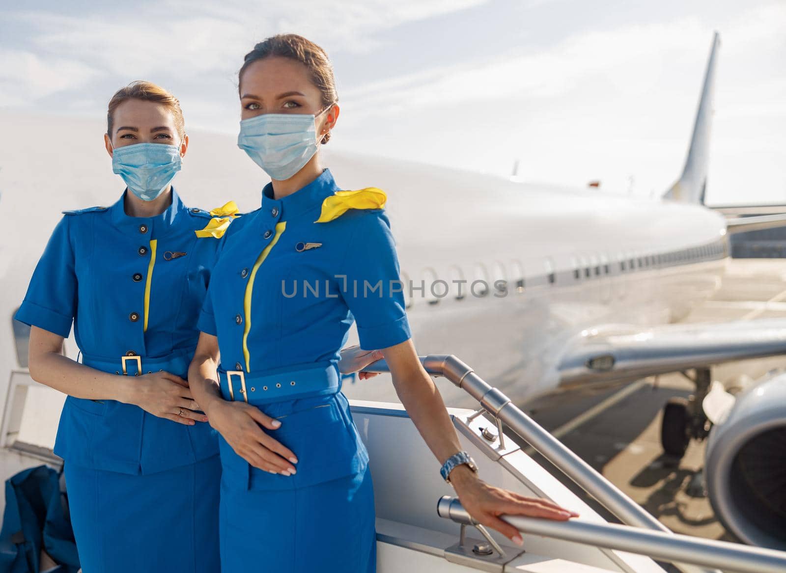 Two beautiful air stewardesses in blue uniform and protective face masks looking at camera, standing on airstair during boarding by Yaroslav_astakhov