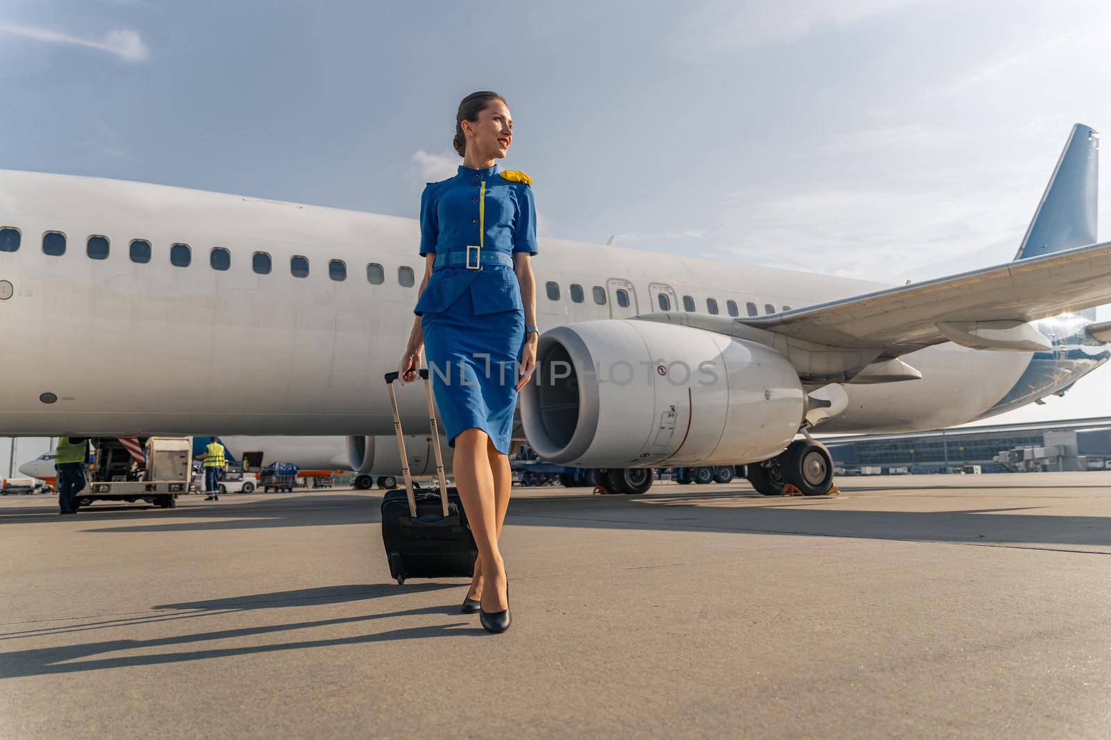 Happy stewardess holding black suitcase and walking on runway with airplane in the background