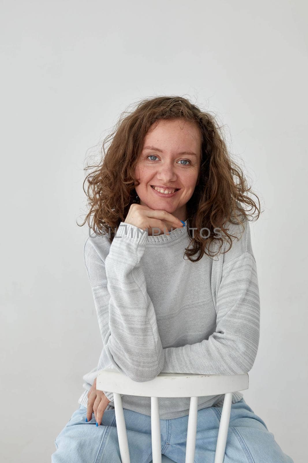 Charming young female with wavy hair dressed in casual gray sweater and jeans sitting on chair and looking at camera with smile against white background