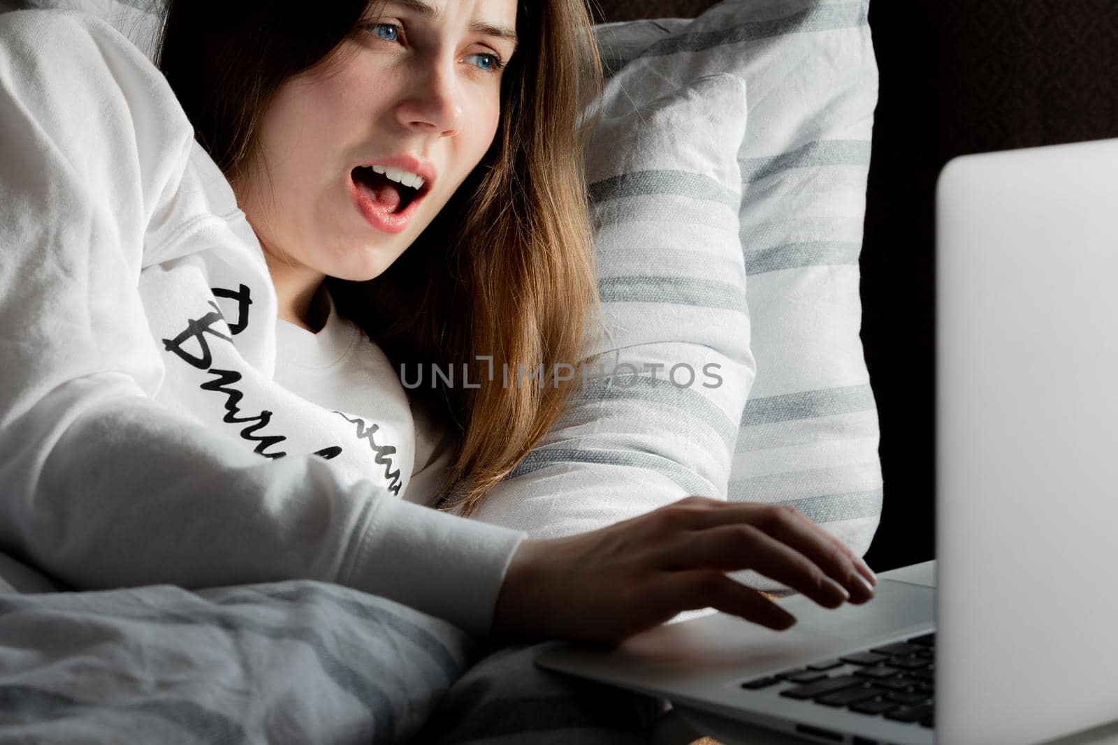 A young cute girl on a sunny day in a white jacket lying on the bed and looking at the laptop in surprise.