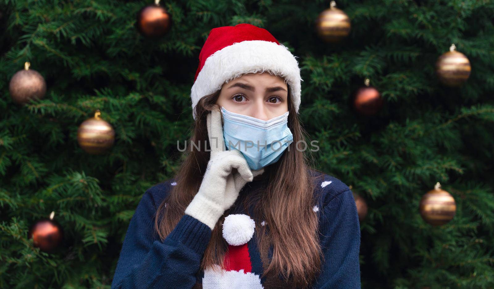 Choosing a gift. Close up Portrait of woman wearing a santa claus hat and medical mask with emotion. Against the background of a Christmas tree. Coronavirus pandemic
