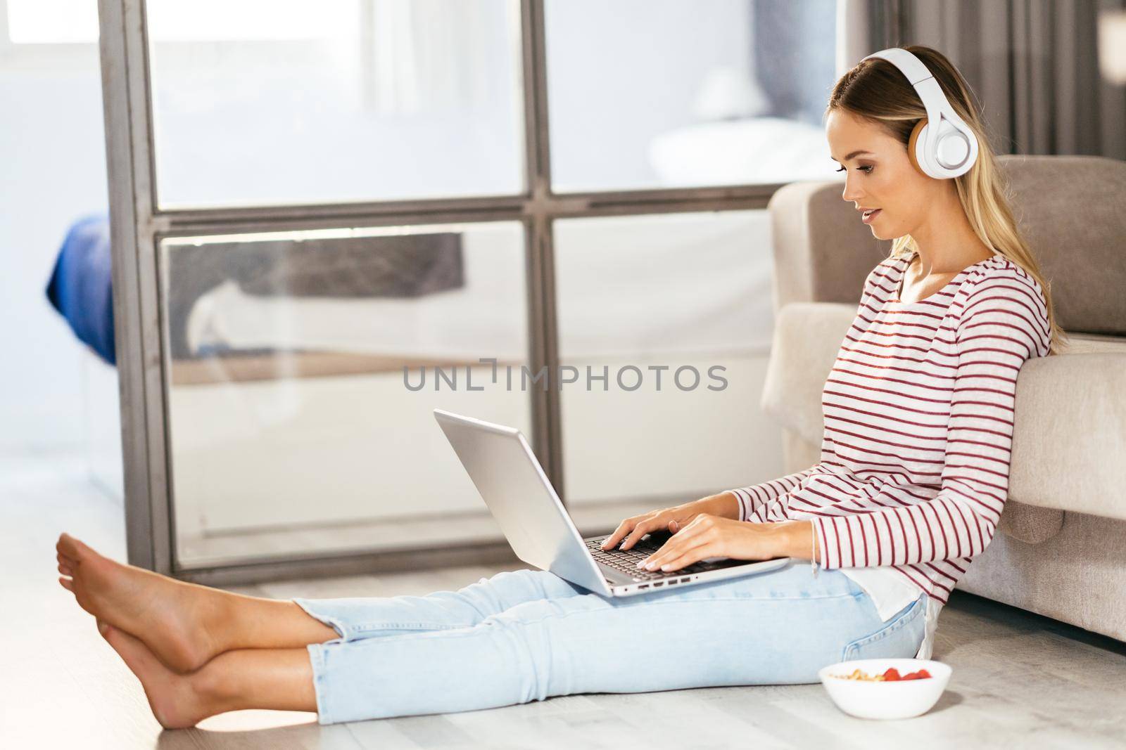 Caucasian blond woman with headphones and laptop on the sofa at home.