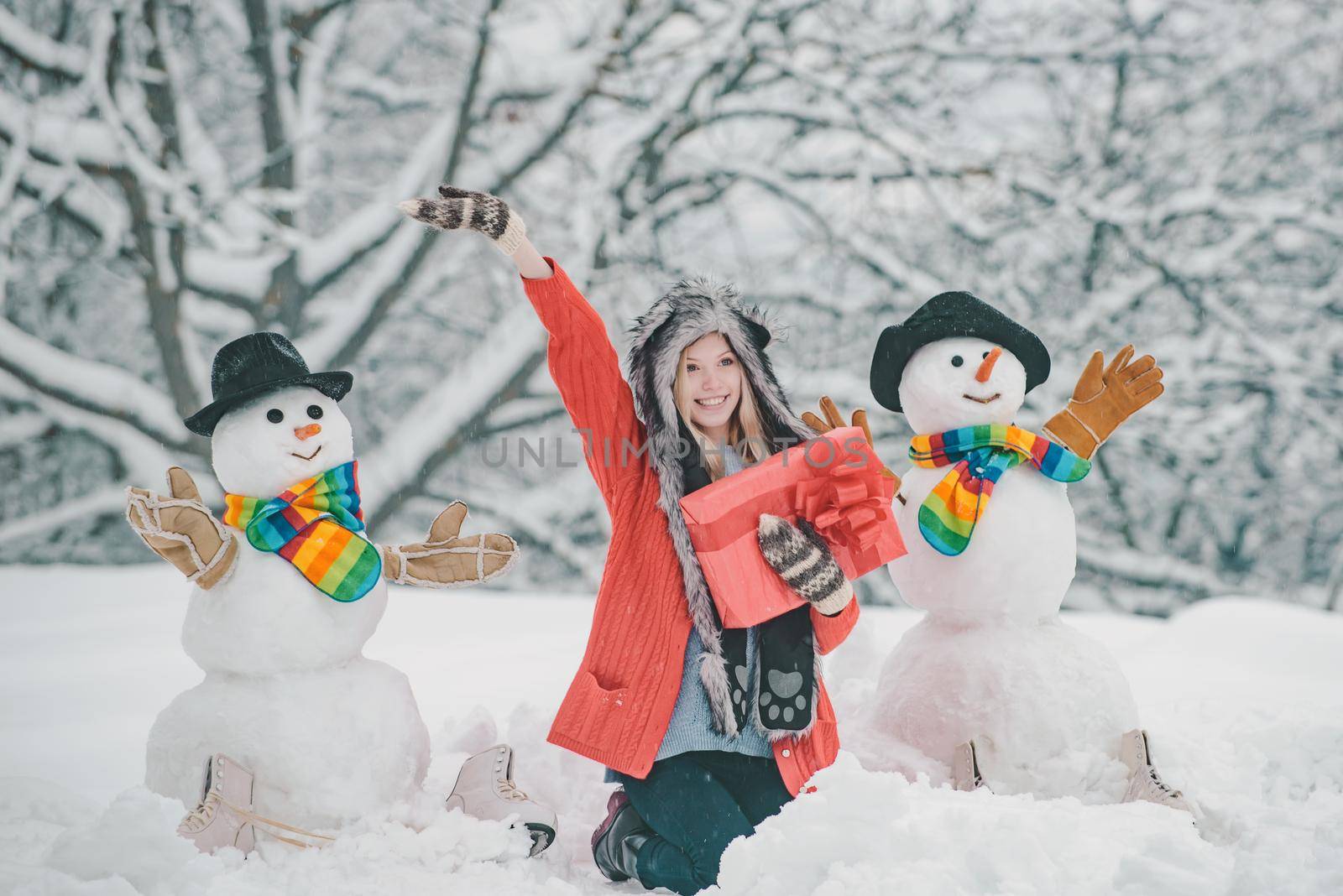 Girl playing with snowman in winter park. Snowman and funny young woman in winter hat and scarf. by Tverdokhlib