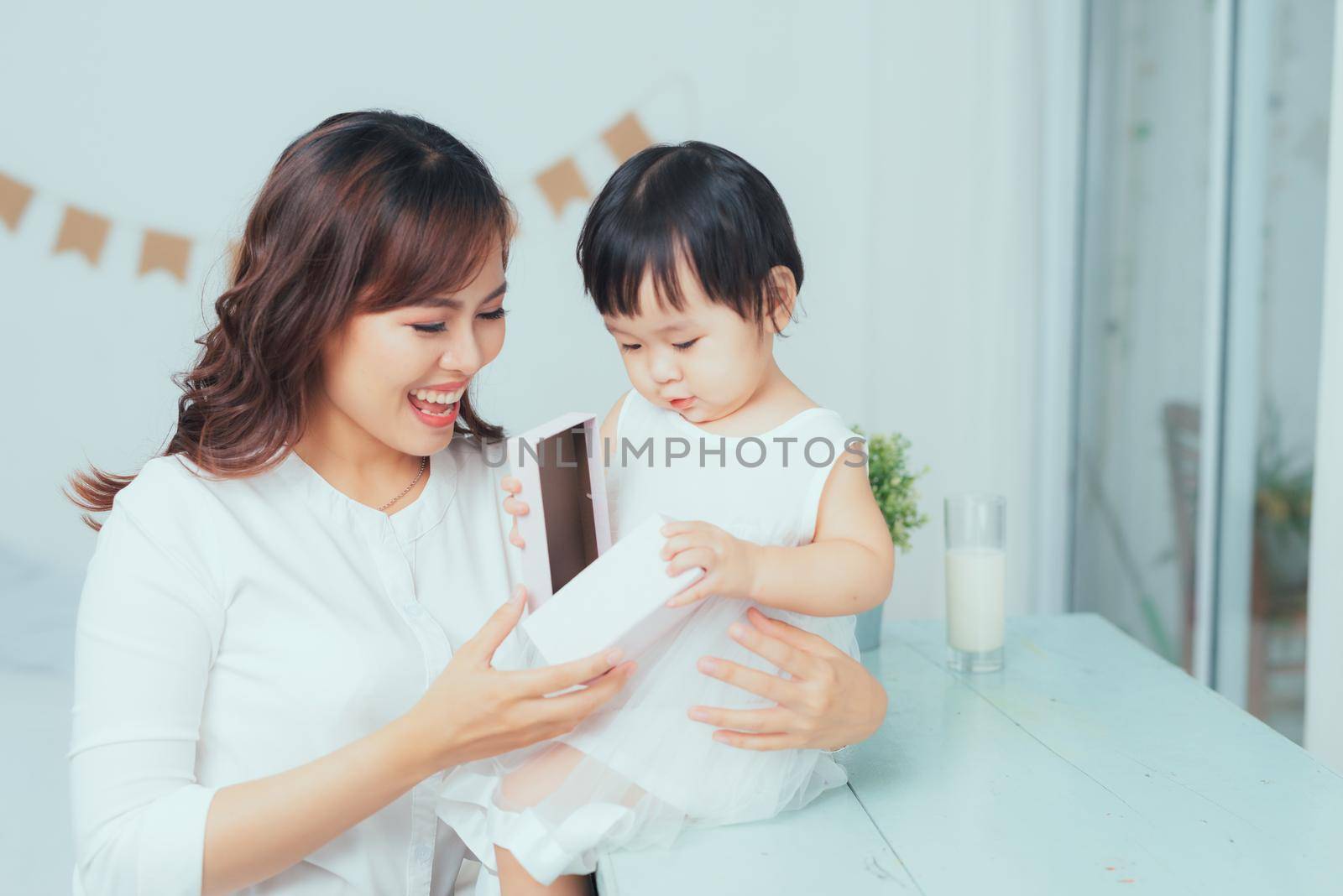 Beautiful young mother smiling while her baby daughter opening birthday gift box. by makidotvn