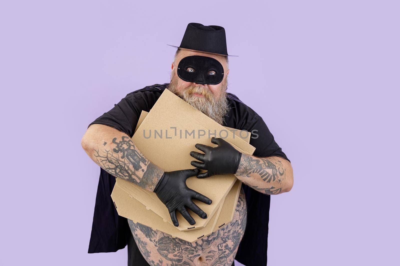 Greedy mature man in Zorro costume with overweight and tattoos hugs brown boxes of pizza on purple background in studio