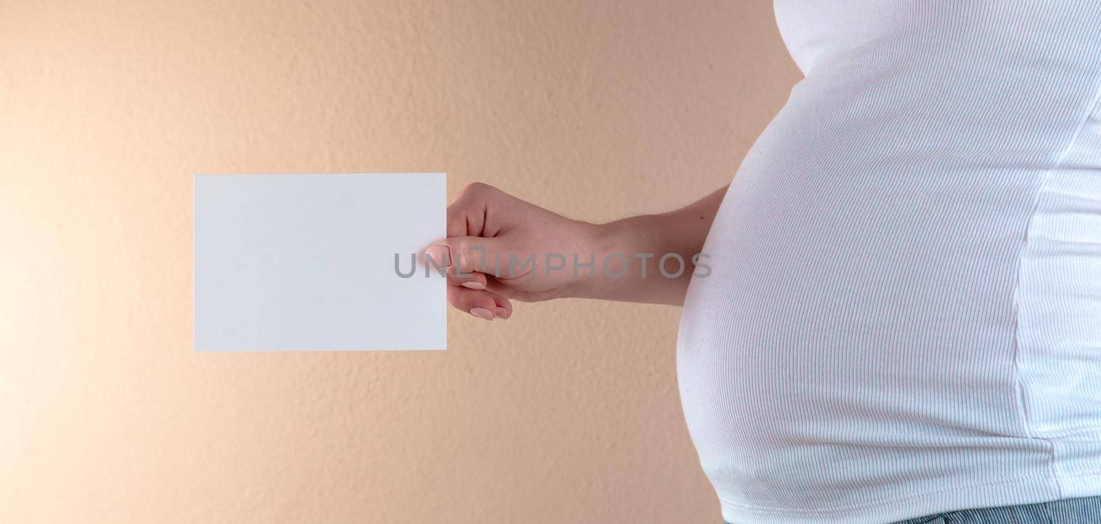 A close-up view of the belly of a pregnant woman in a white T-shirt that is holding an empty sheet of paper