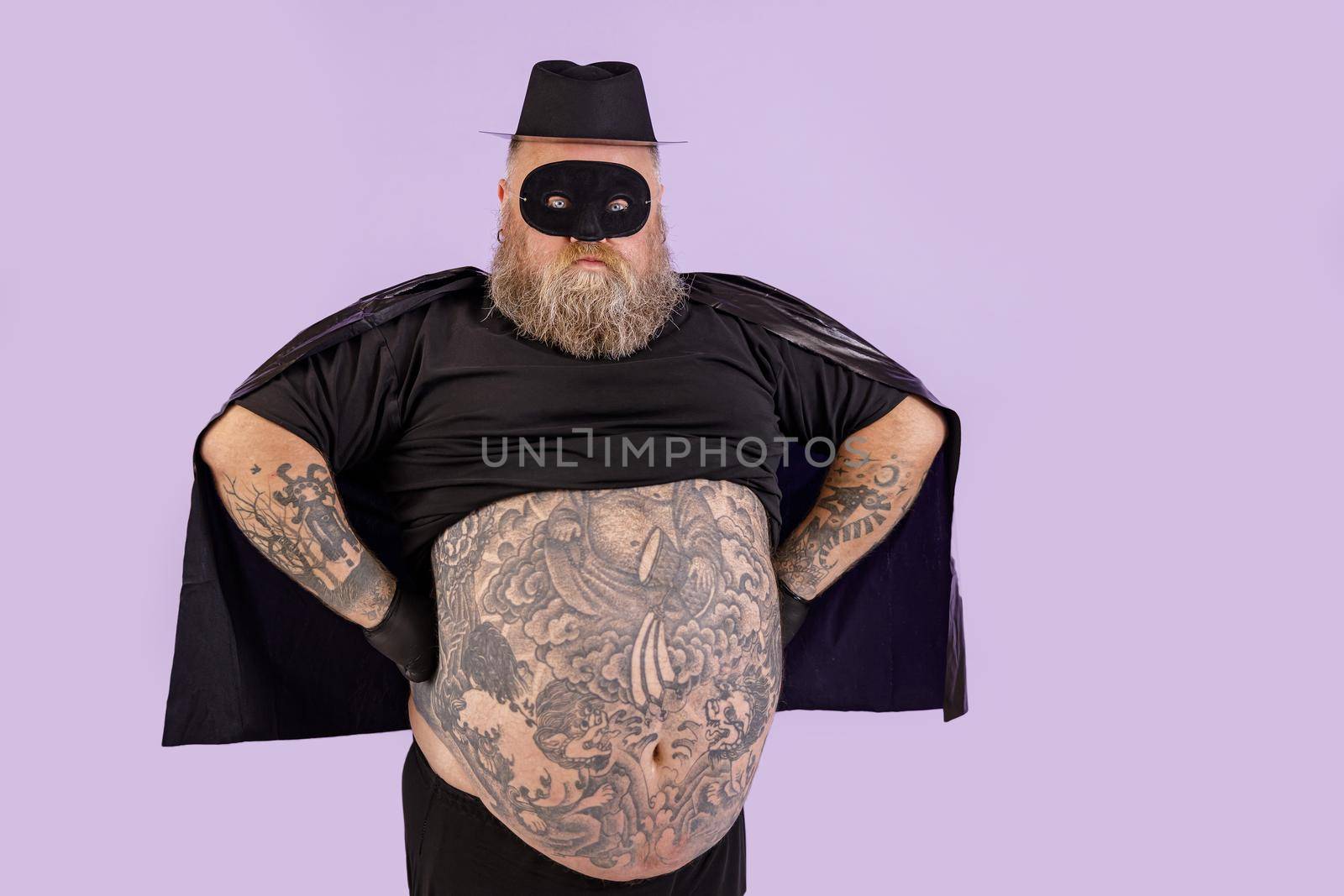 Confident man with overweight in Zorro suit holds hands on waist on purple background by Yaroslav_astakhov