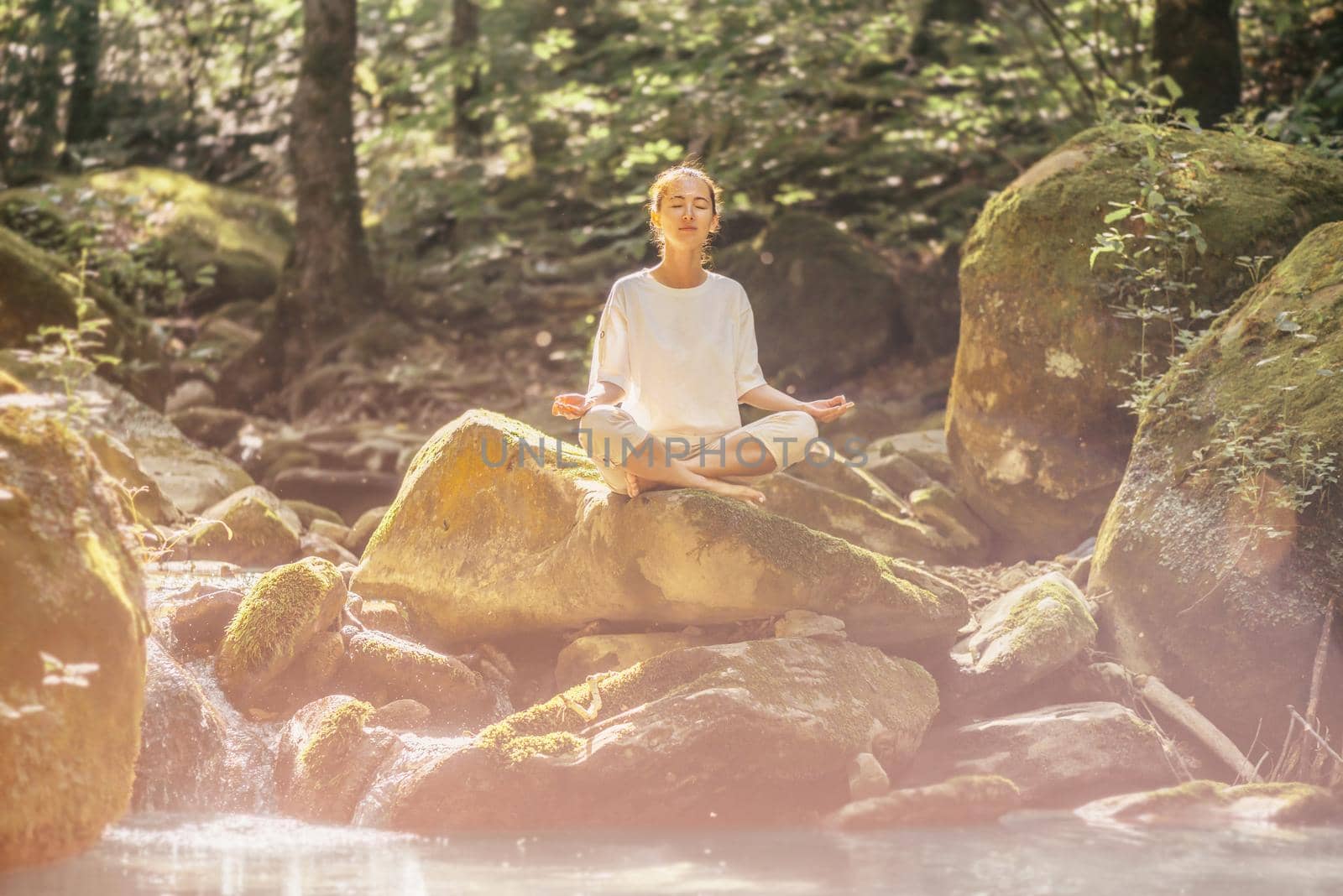 Beautiful young woman practicing yoga with closed eyes in pose of lotus on stone near the river in forest outdoor.