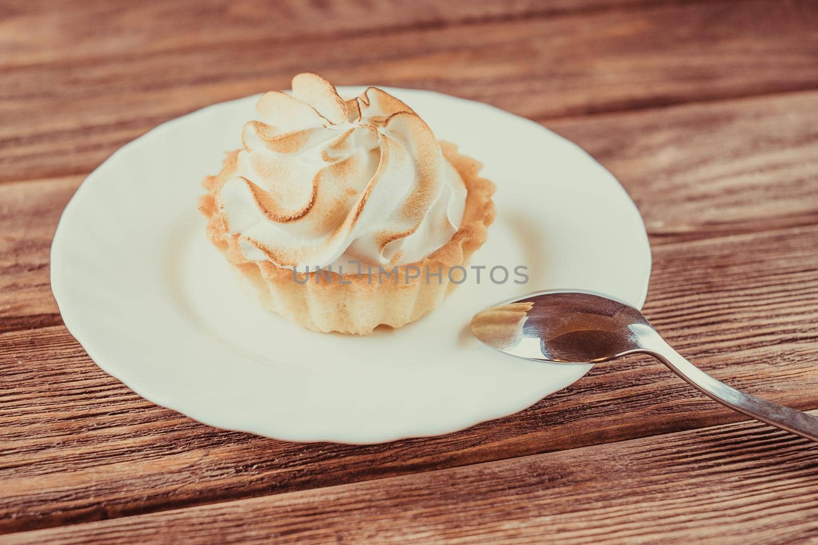 Cupcake with whipped cream on a white saucer with teaspoon on a wooden table.