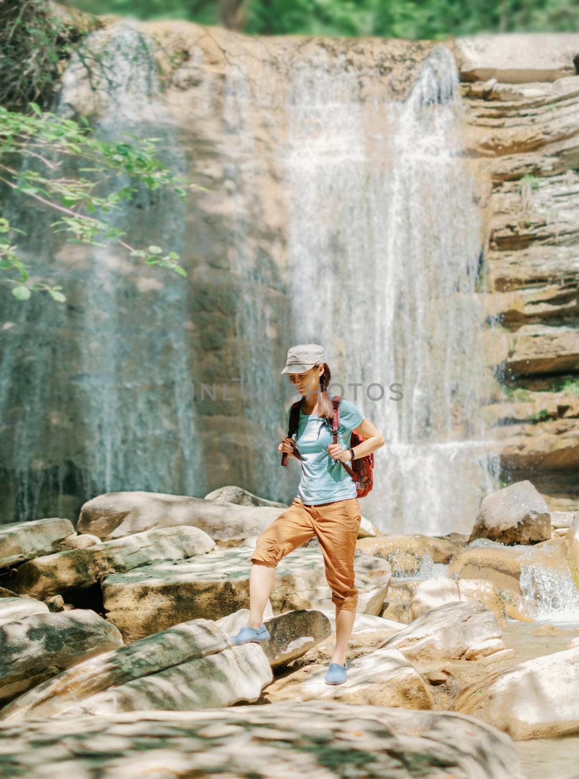 Explorer backpacker young woman walking on stones in front of waterfall outdoor on sunny day.