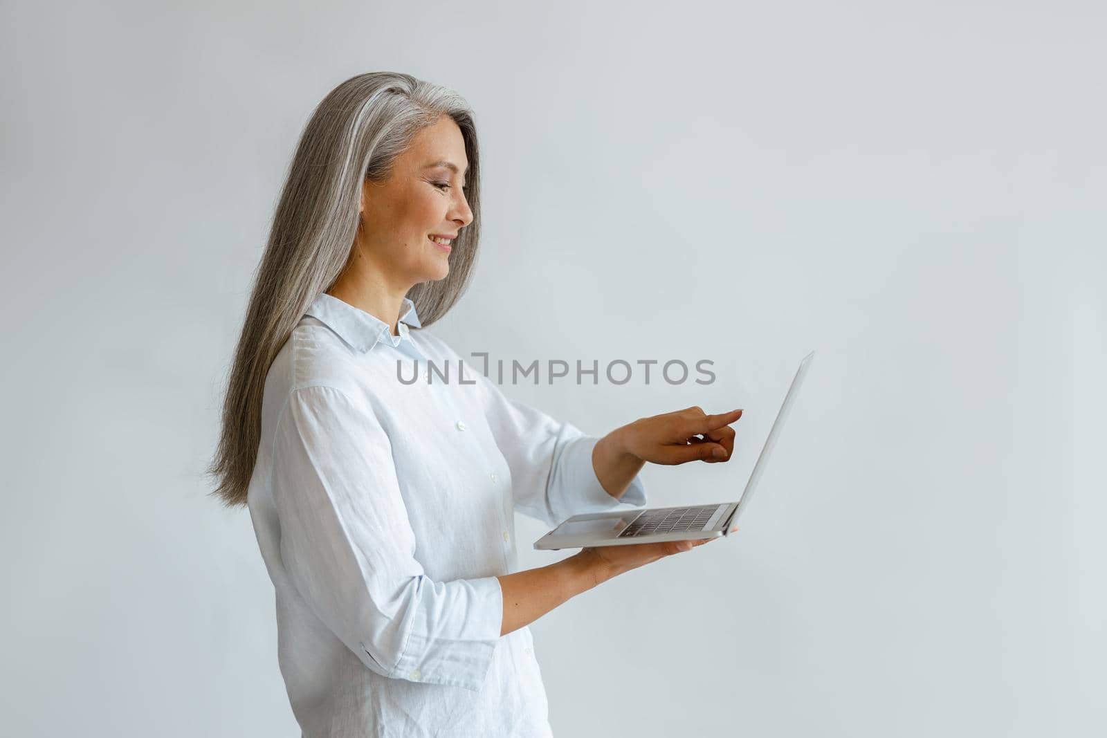 Smiling mature Asian businesswoman points onto screen of laptop on light background by Yaroslav_astakhov