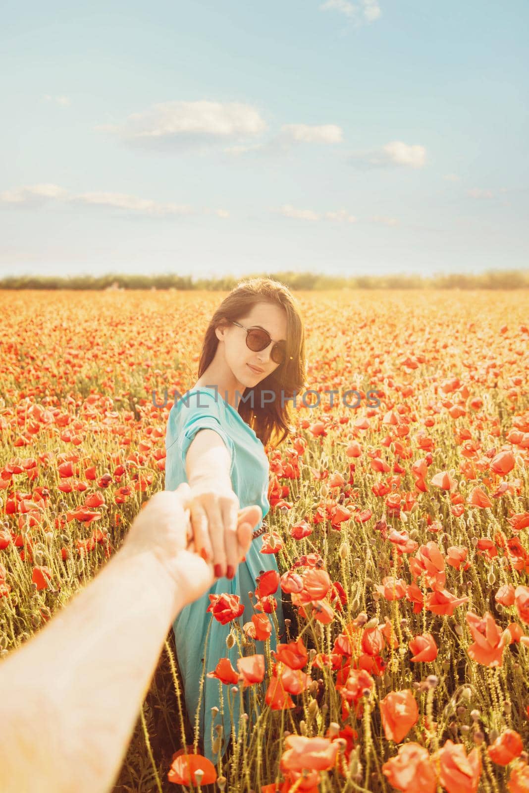 Beautiful young woman holding man's hand and leading him in poppies meadow outdoor, looking at camera. Point of view.