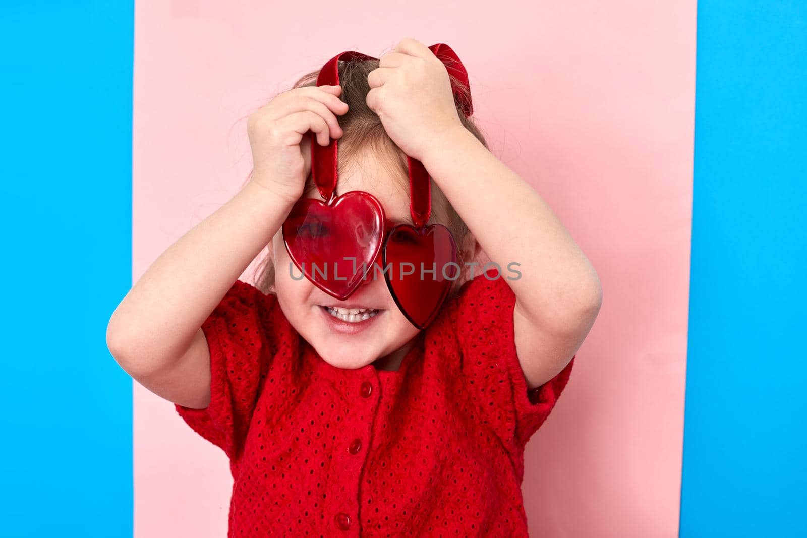 Cute delighted little girl standing in studio and looking at camera through red plastic toy hearts