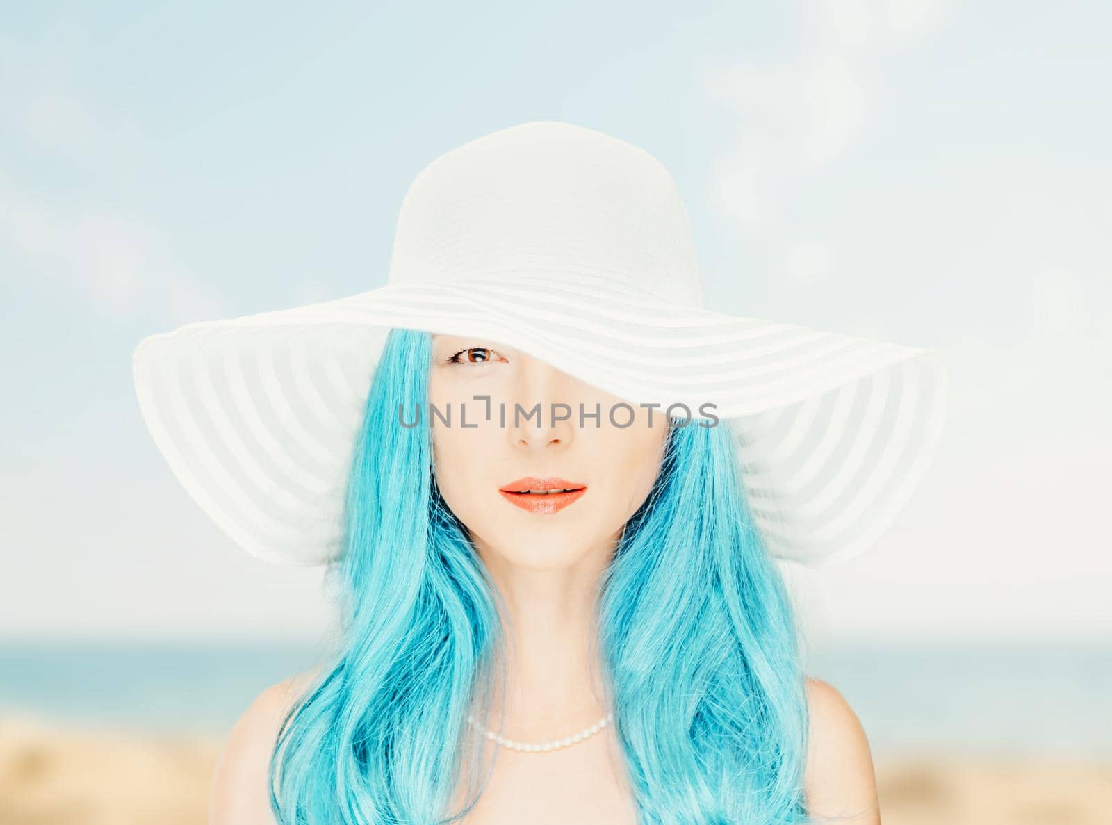 Portrait of stylish attractive woman with blue hair in a white hat with wide brim on background of beach, looking at camera.
