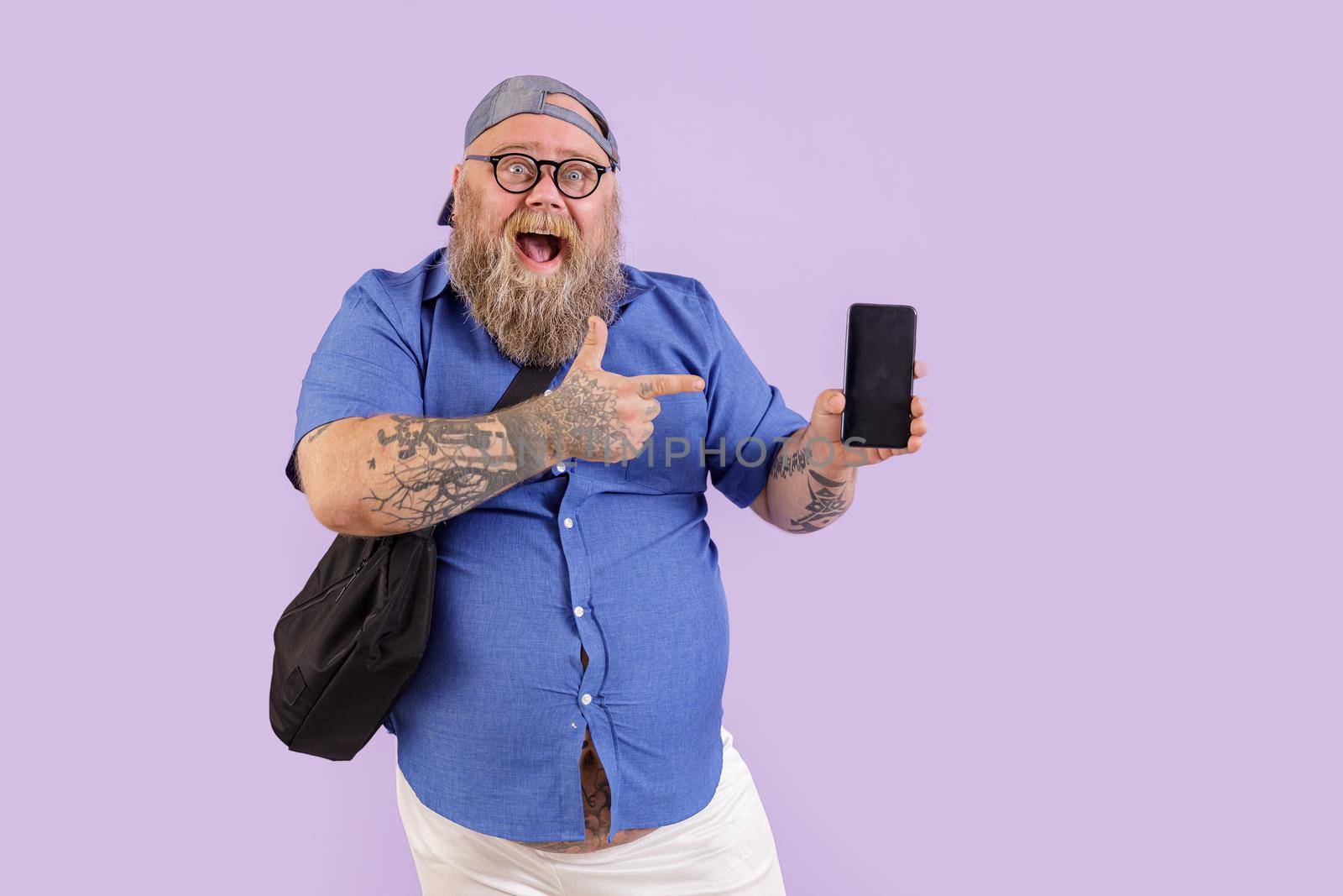 Positive middle aged fat man with crossbody bag points onto mobile phone with blank screen standing on purple background in studio