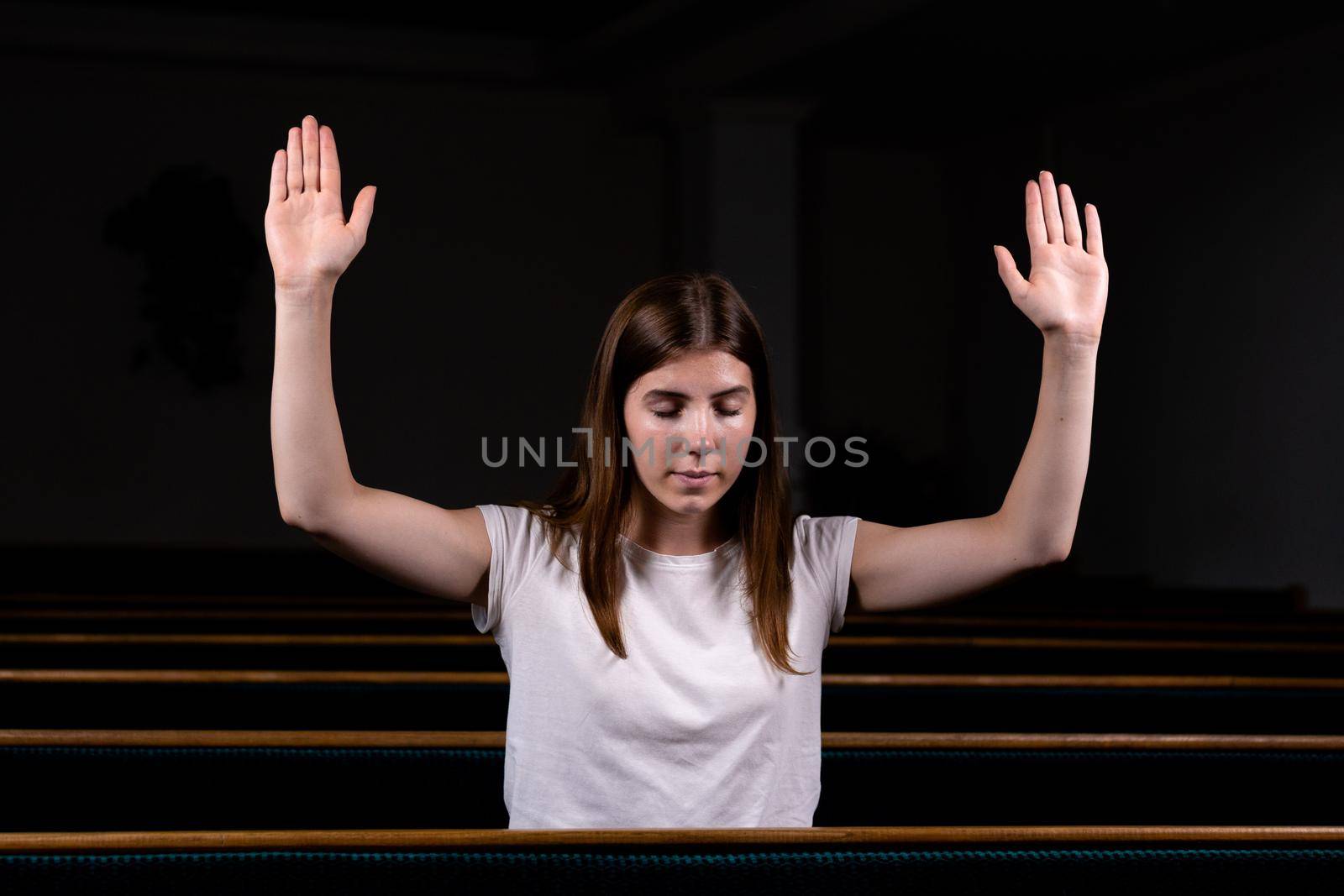 A Christian girl in white shirt is sits with his hands up and face and praying with humble heart in the church by lunarts