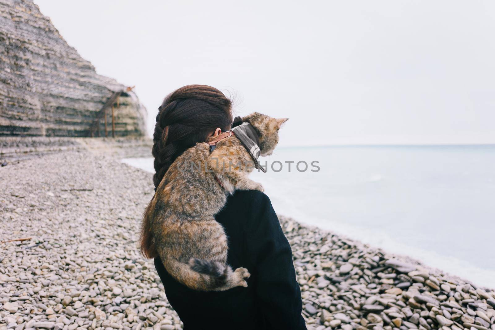 Young woman with cat on her shoulder standing on pebble coastline near the sea.