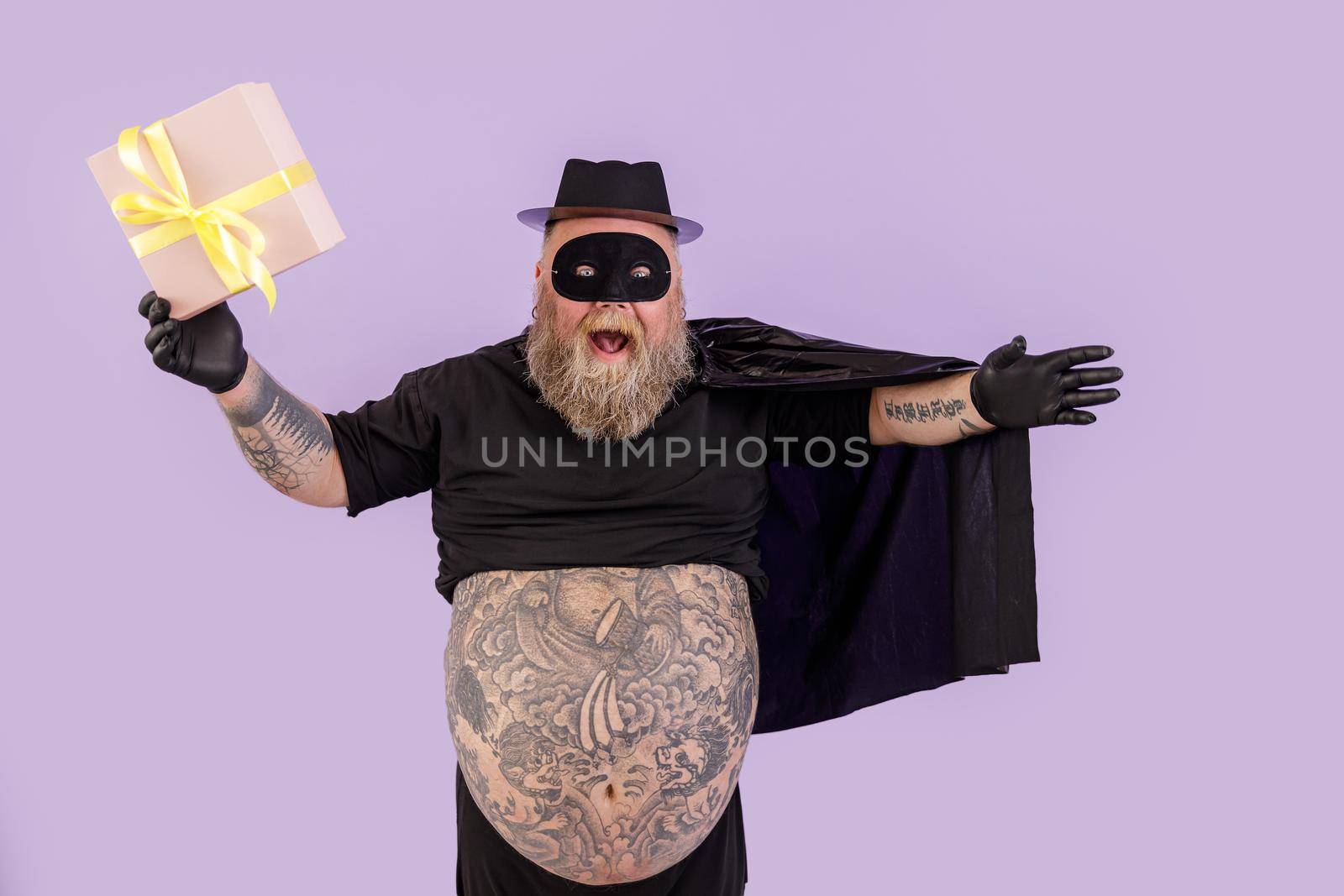 Joyful man with overweight in hero costume with cape holds gift on purple background by Yaroslav_astakhov