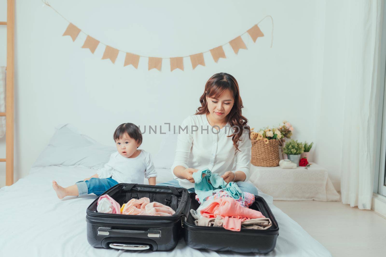 cute baby sits beside the mother preparing clothes to put in the suitcase