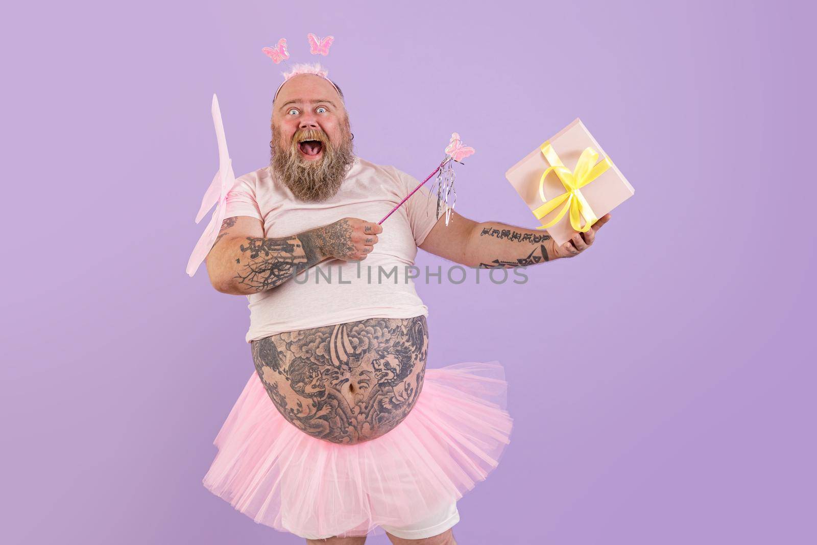 Excited obese male person in fairy costume holds gift box on purple background by Yaroslav_astakhov