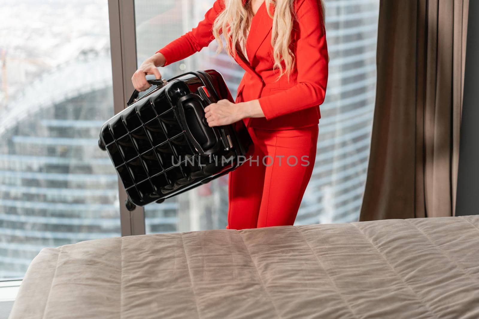 Stewardess in red uniform arrives in a hotel room with black suitcase. Rest in the transit city before the return flight. Young blond Woman in red coral suit unpacks suitcase