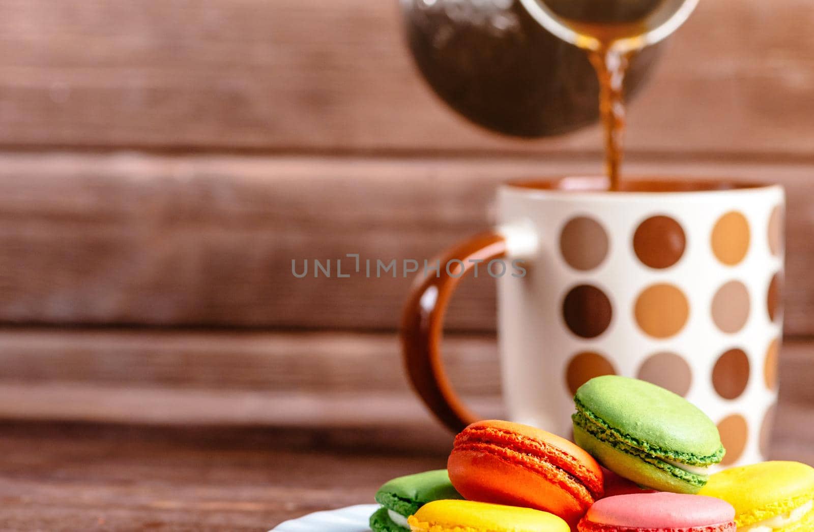 Pouring hot coffee from cezve to cup near sweet macaroons dessert, focus on foreground.