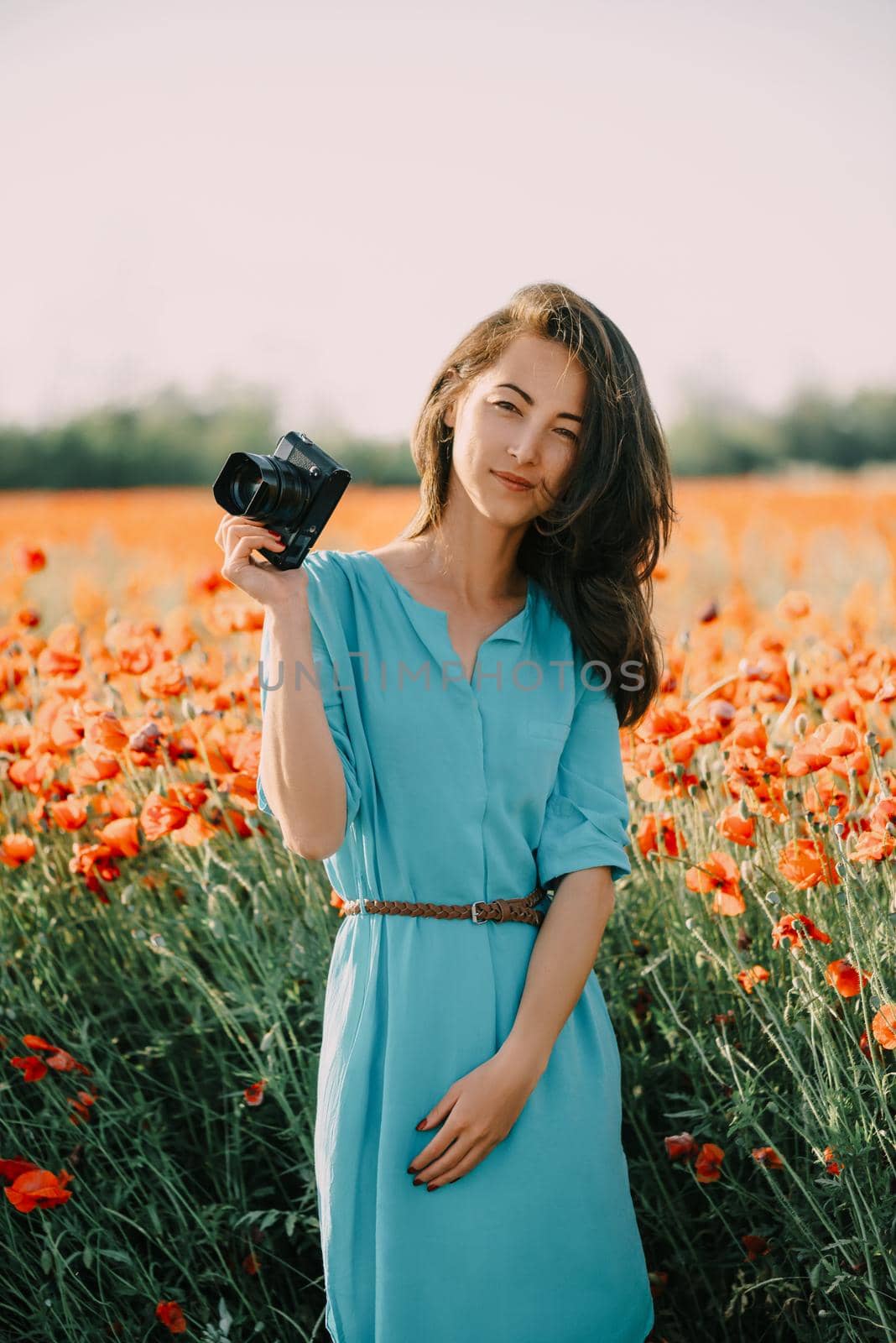 Beautiful brunette girl in blue dress with photo camera standing in poppy meadow, looking at camera.