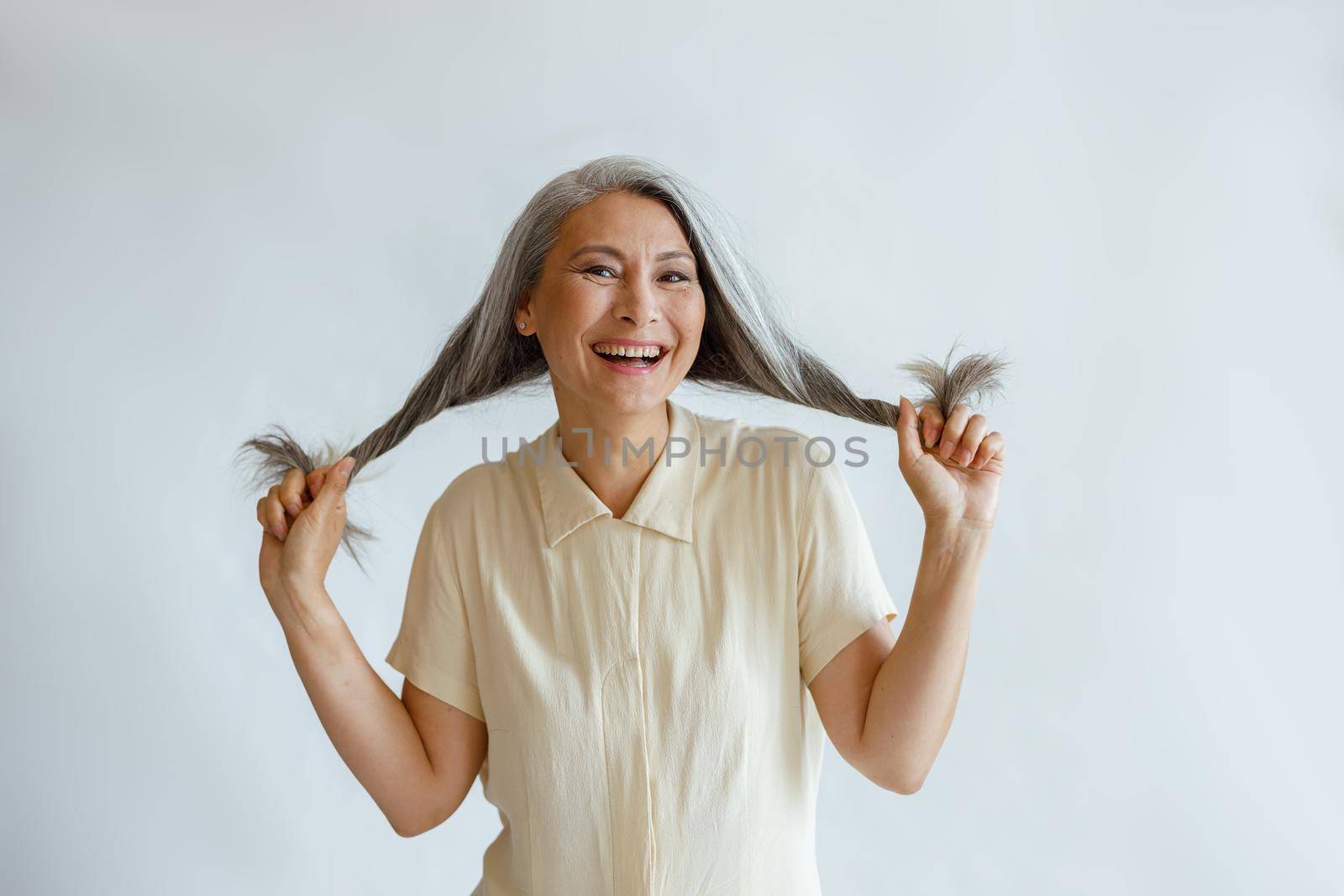 Happy middle aged Asian woman holds twisted hoary hair on light grey background by Yaroslav_astakhov