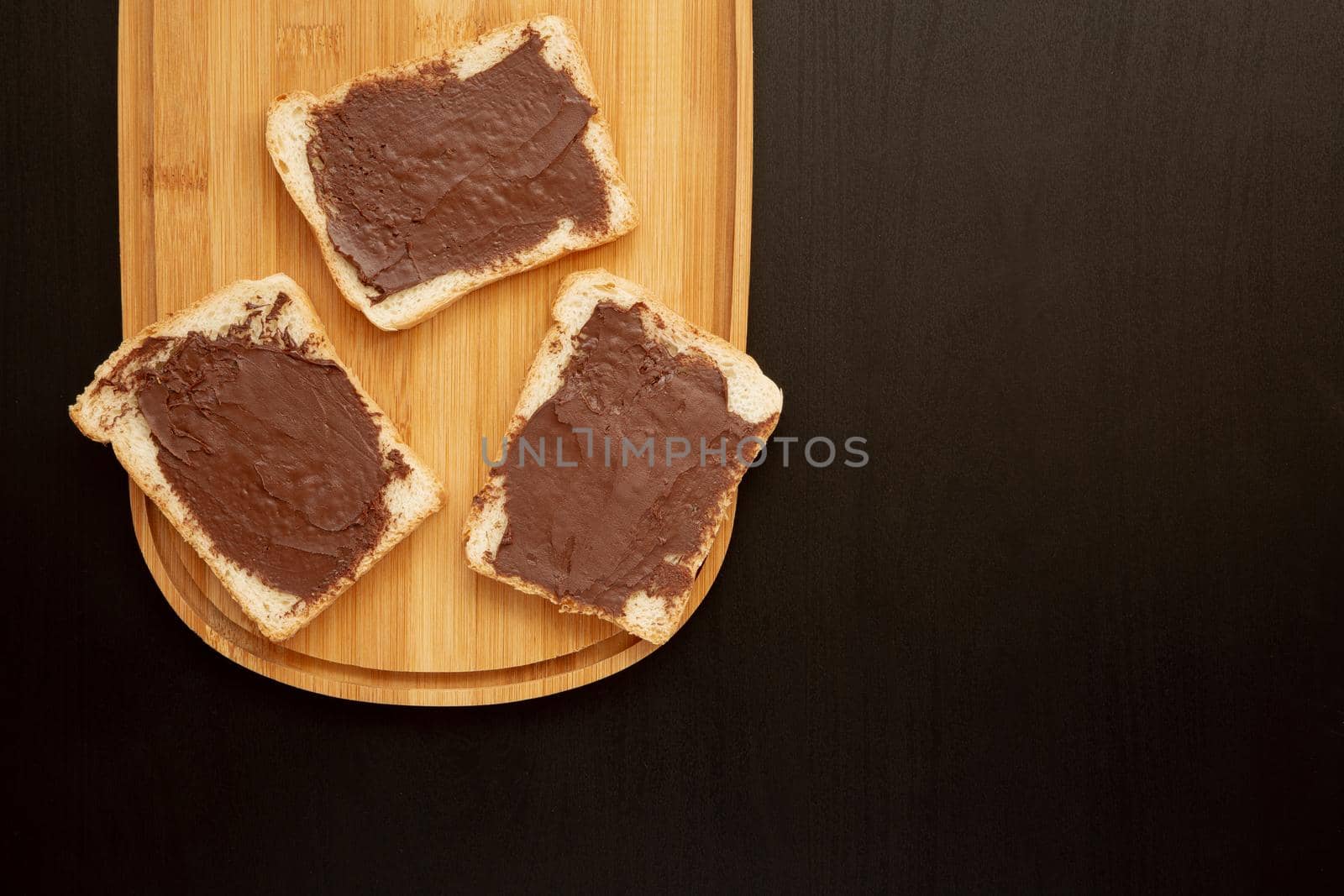 A composition of three white toasts smeared with chocolate butter that lie on a cutting board against a dark background. top view with area for text