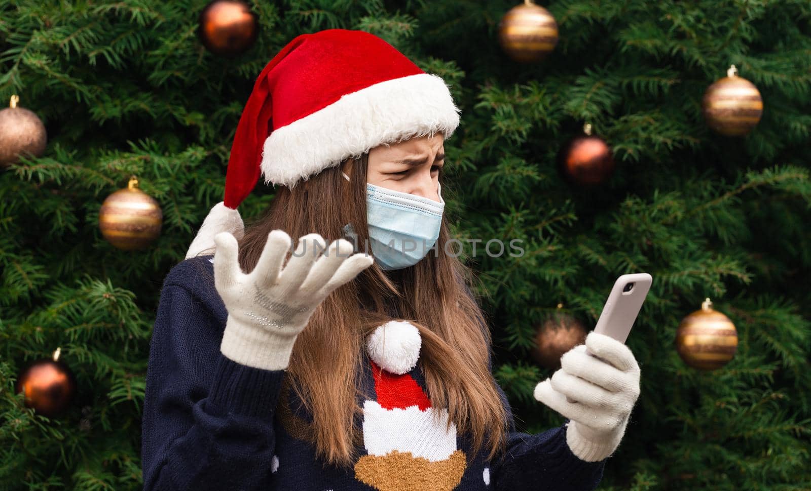 Christmas online greetings. Close up Portrait of woman wearing a santa claus hat and medical mask with emotion. Against the background of a Christmas tree. Coronavirus pandemic by lunarts