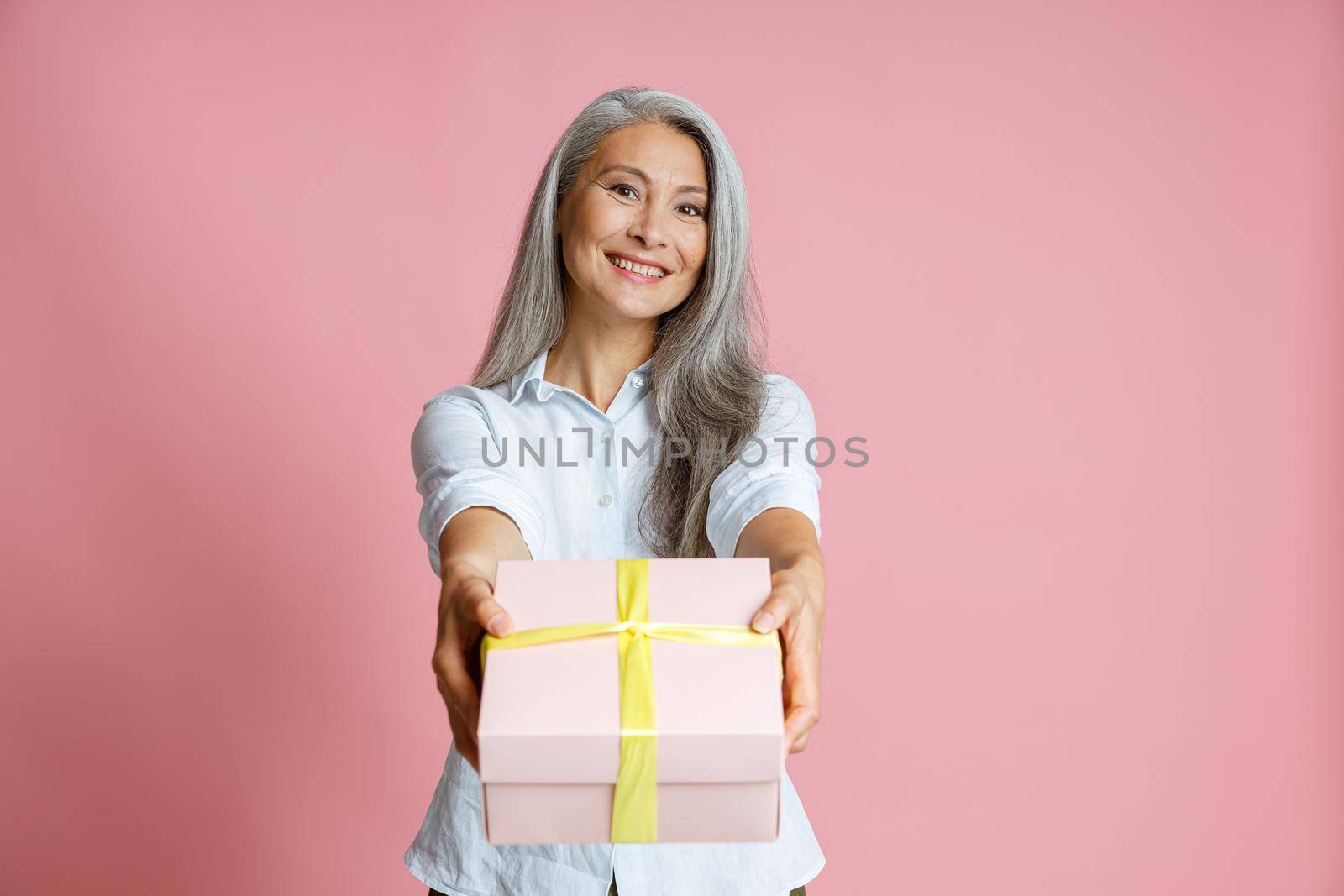 Pretty middle aged Asian lady with long loose hoary hair presents gift decorated with ribbon standing on pink background in studio
