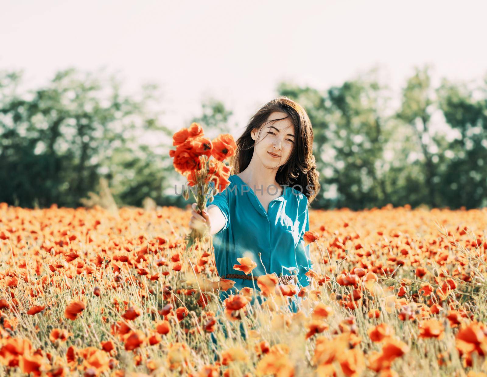 Smiling beautiful young woman standing with bouquet of poppies in flower field, looking at camera.