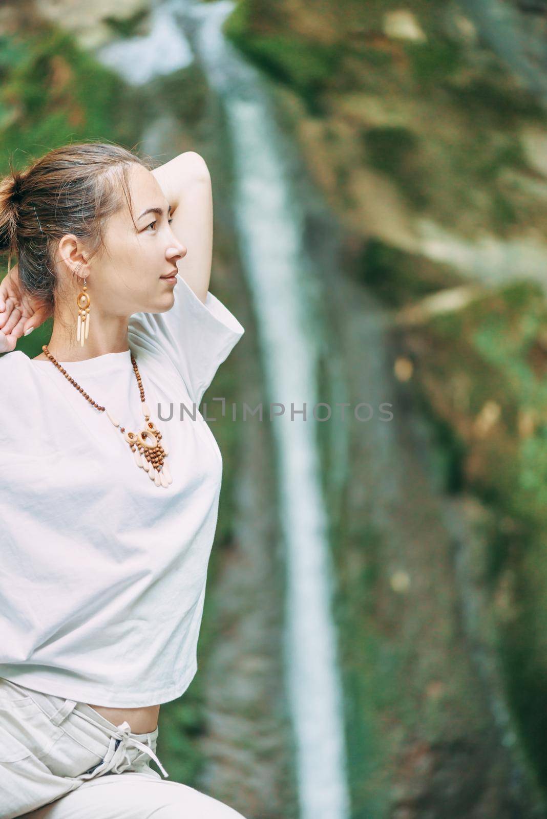 Smiling beautiful young woman practicing yoga in front of waterfall outdoor.