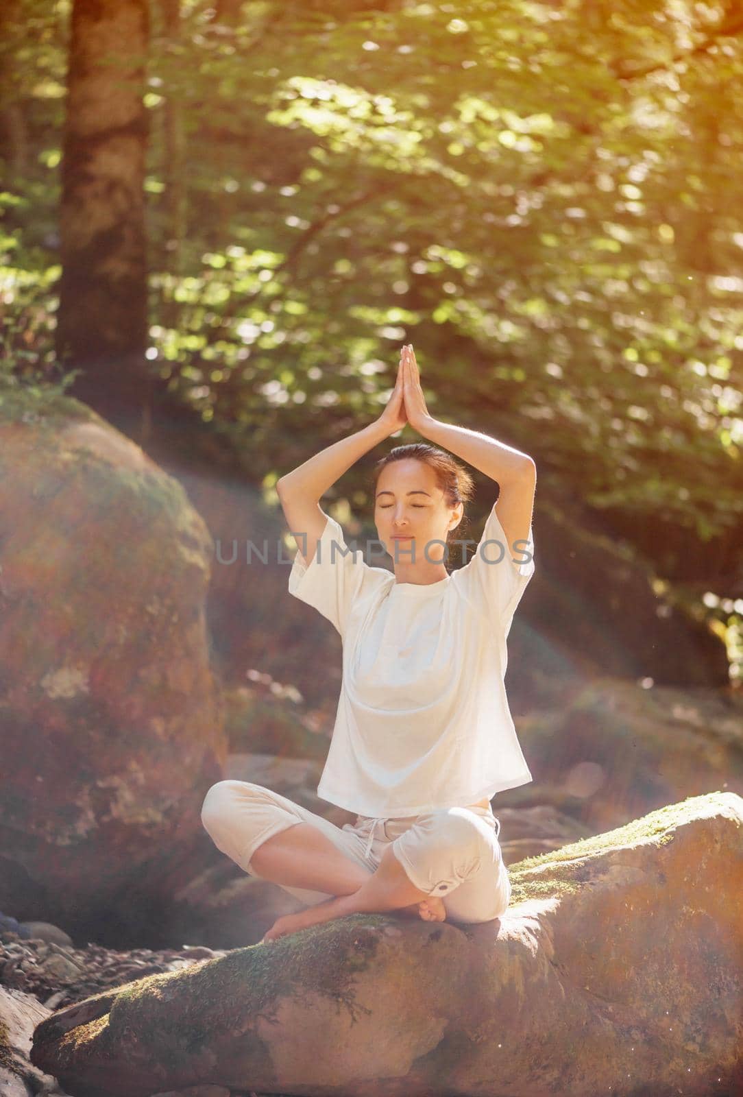 Beautiful young woman meditating with closed eyes in pose of lotus on stone in forest outdoor.