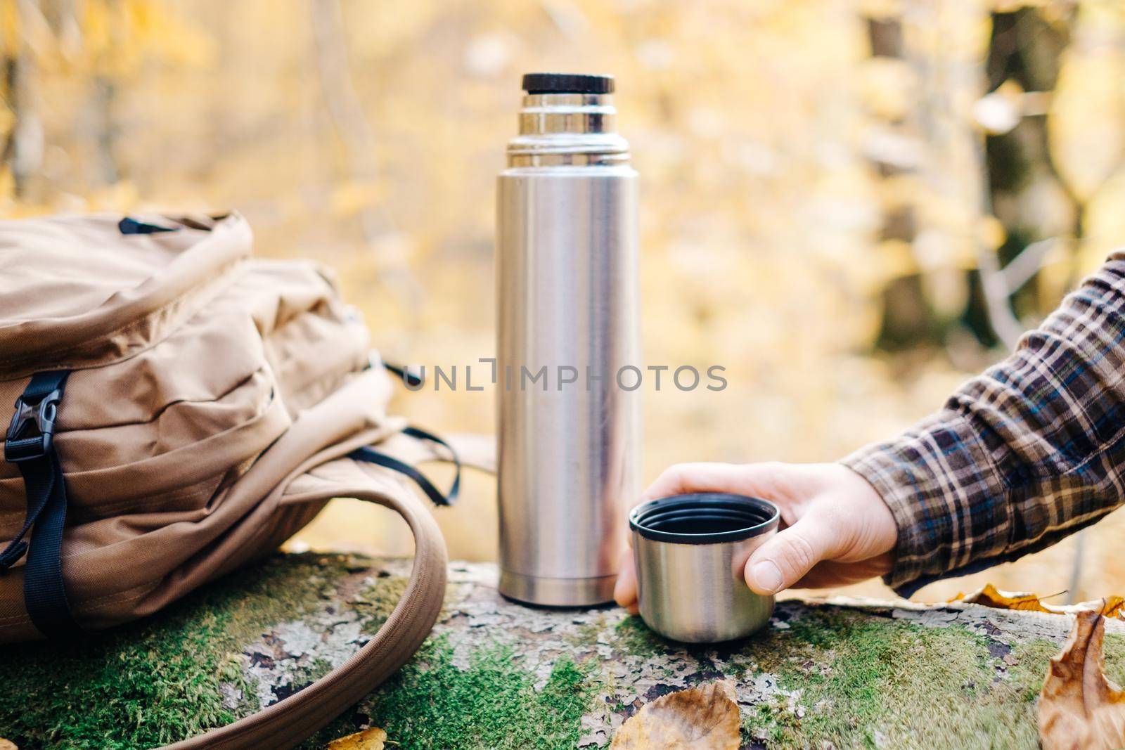 Male hand holding cup near the thermos and backpack in autumn forest.