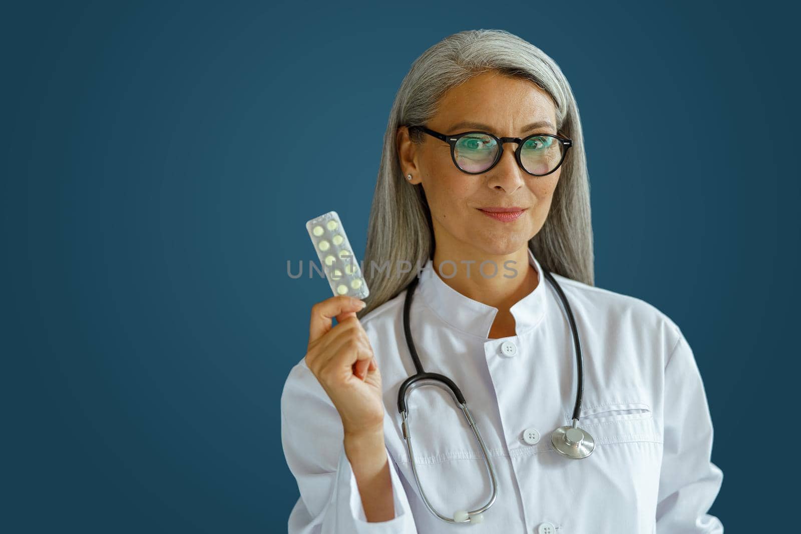 Smiling middle aged female therapist in white uniform holds pills blister standing on blue background by Yaroslav_astakhov