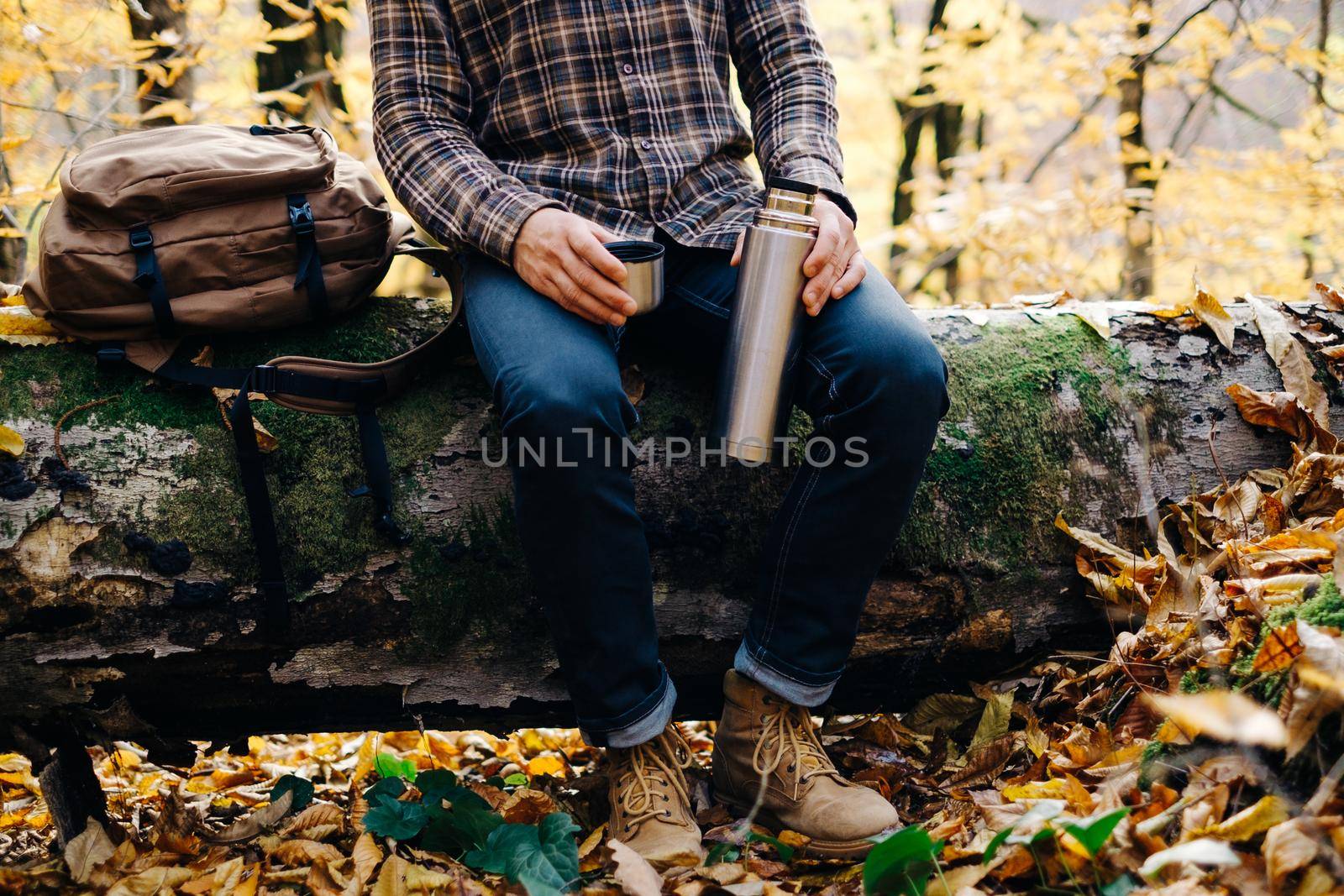 Unrecognizable backpacker young man sitting on fallen tree trunk with cup and thermos and resting in autumn forest.