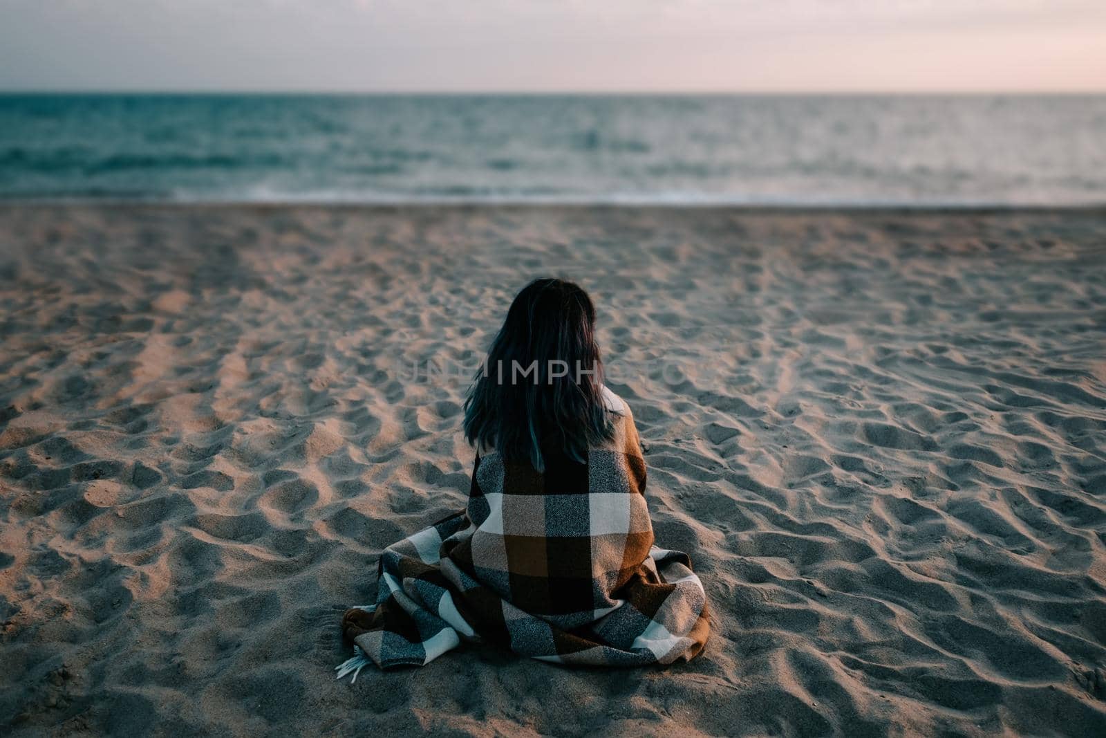 Young woman wrapped in plaid sitting on sand beach in front of sea on vacations.