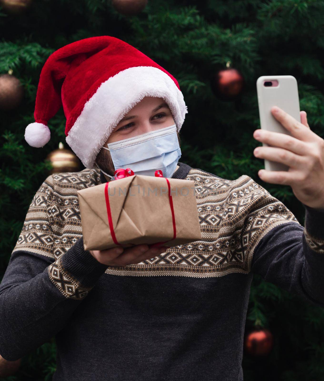 Christmas online congratulations. Portrait man wearing santa hat and medical mask, giving gift present box with red ribbon, christmas tree bokeh on background