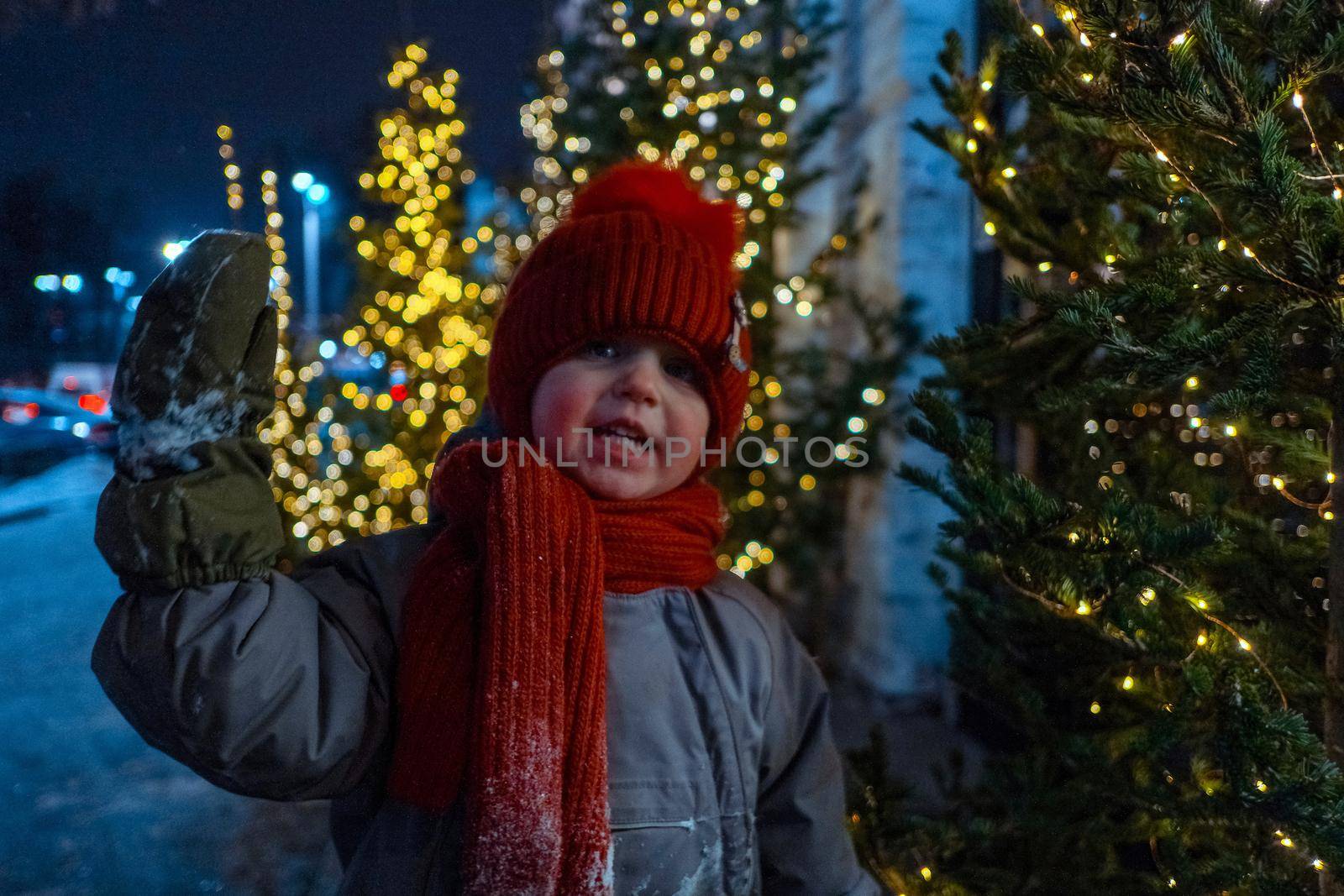 Happy child in warm outerwear waving hand and looking at camera while standing near spruce tree decorated with glowing lights in evening city during Christmas holidays