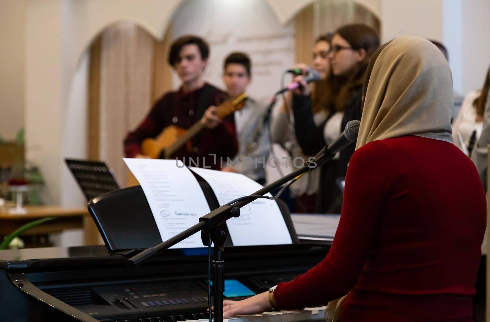 KOROSTEN - NOV, 10, 2019: In the foreground is a view from the back of a girl who plays the piano in a modern church. A music band is in blur by lunarts