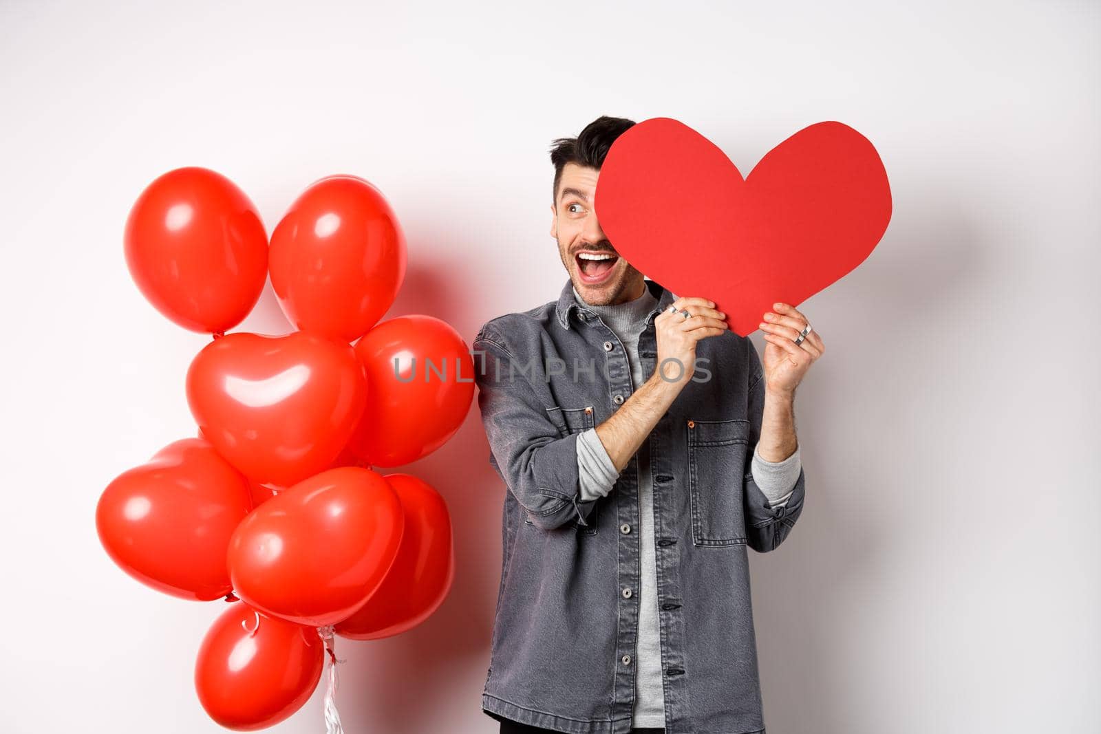 Romantic cheerful man cover half of face with Valentine heart cutout and smiling amazed, celebrating love holiday, standing on white background.