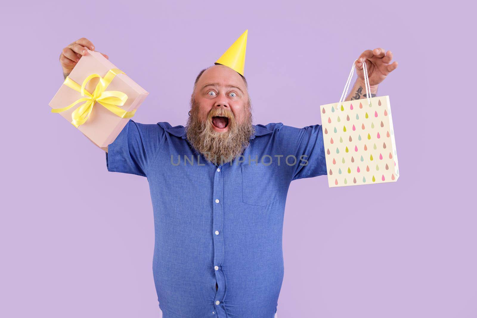 Joyful middle aged bearded obese man in yellow party hat shows gift box and paper bag posing on purple background in studio