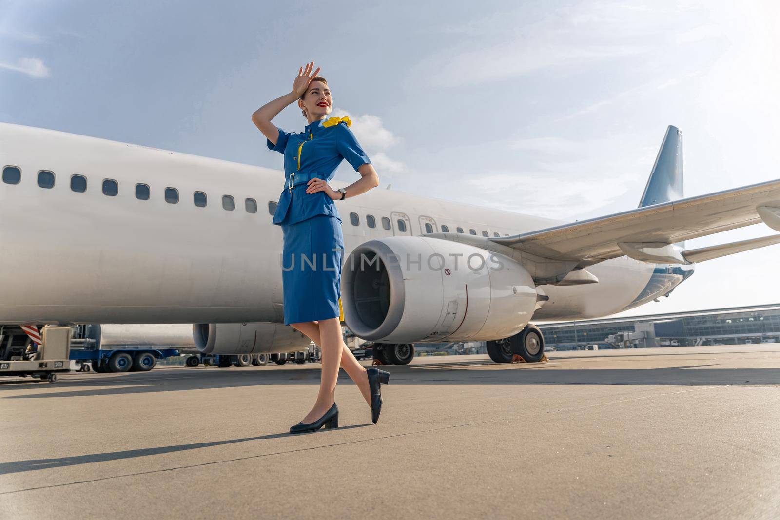 Smiling stewardess in elegant uniform standing on runway with airplane in the background