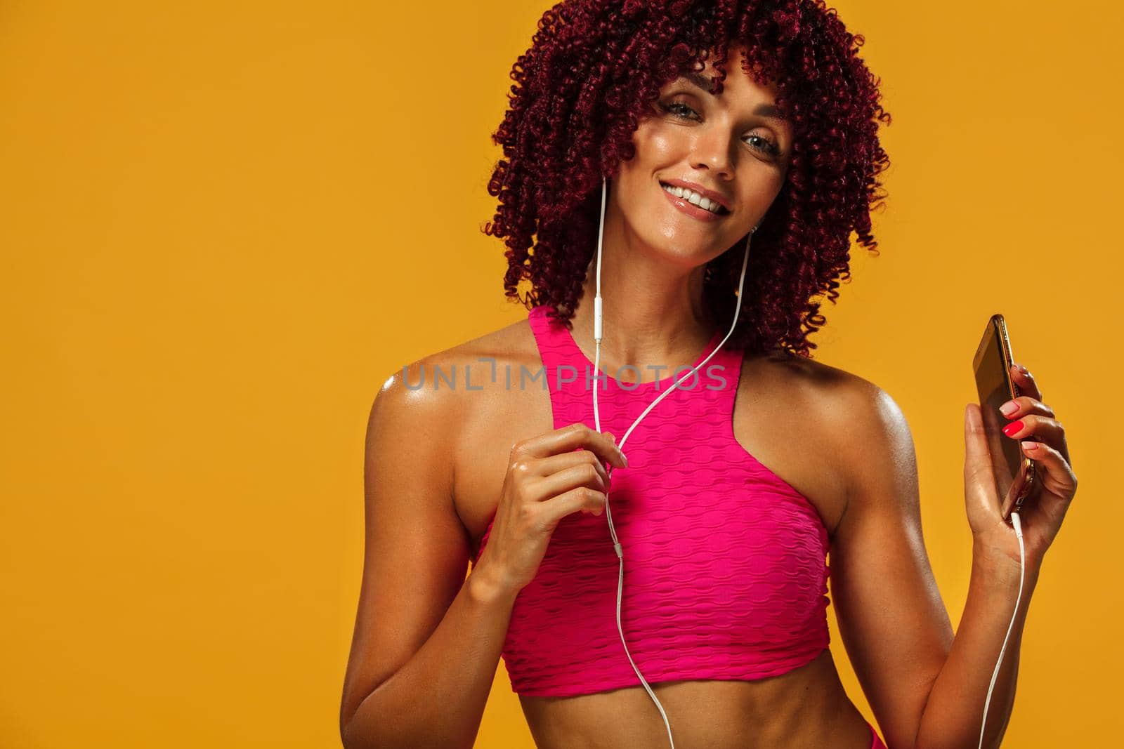 Beautiful young and fit woman in headphones listening music from phone and smiling. Isolated on a yellow background in a pink sportswear by MikeOrlov